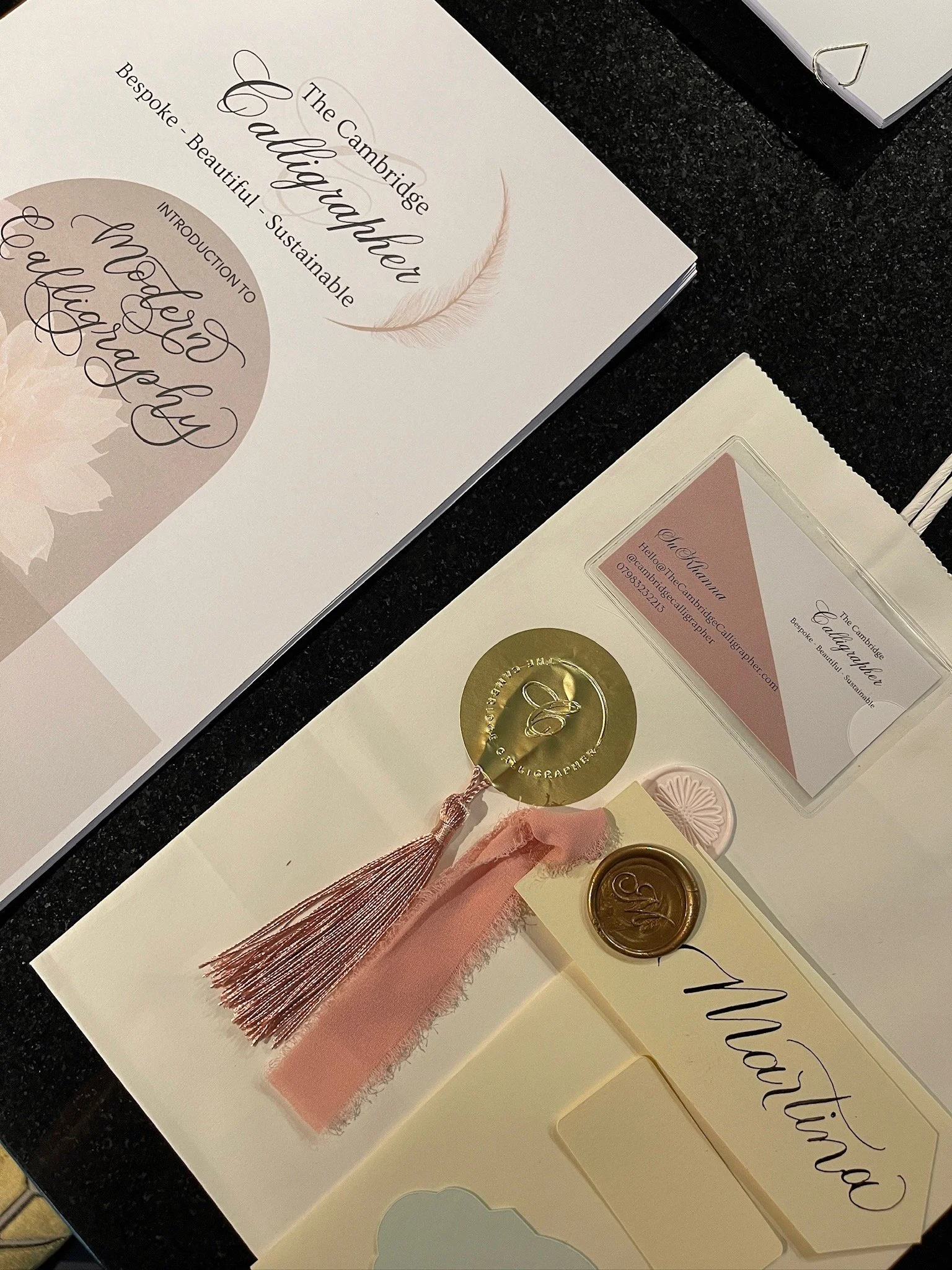 Wedding stationery and calligraphy supplies displayed on a desk, including a booklet titled 'The Cambridge Calligraphy', a name card, wax seals, and decorative embellishments in pink and gold tones.