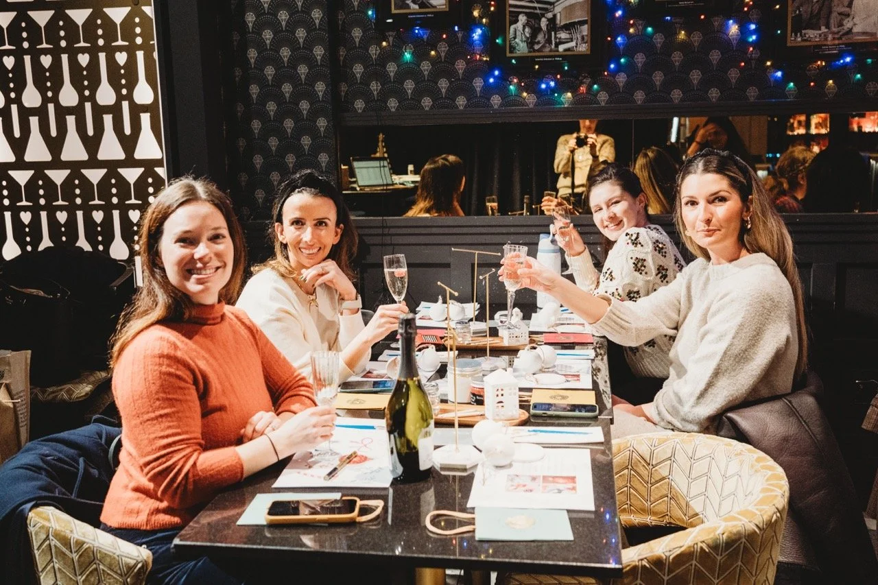 Four women sitting at a restaurant table celebrating with drinks, smiling and toasting. The table has champagne glasses, a bottle of wine, and decorative elements, with a black wall featuring framed photos and colorful string lights in the background