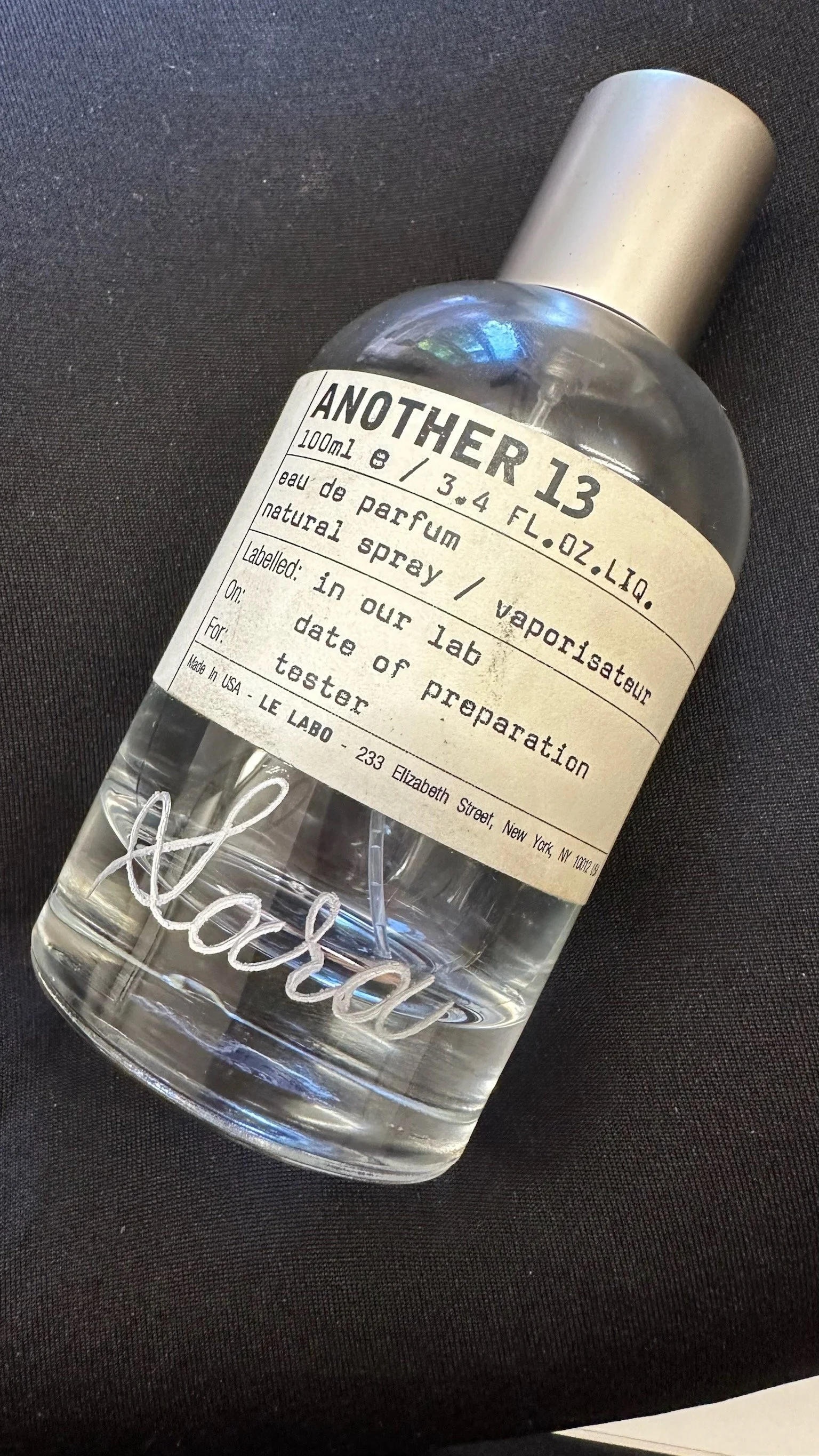 A clear glass spray bottle labeled 'Another 13' containing perfume, with a vintage-style label describing it as a natural spray Eau de parfum, made in the USA, resting on a black surface.