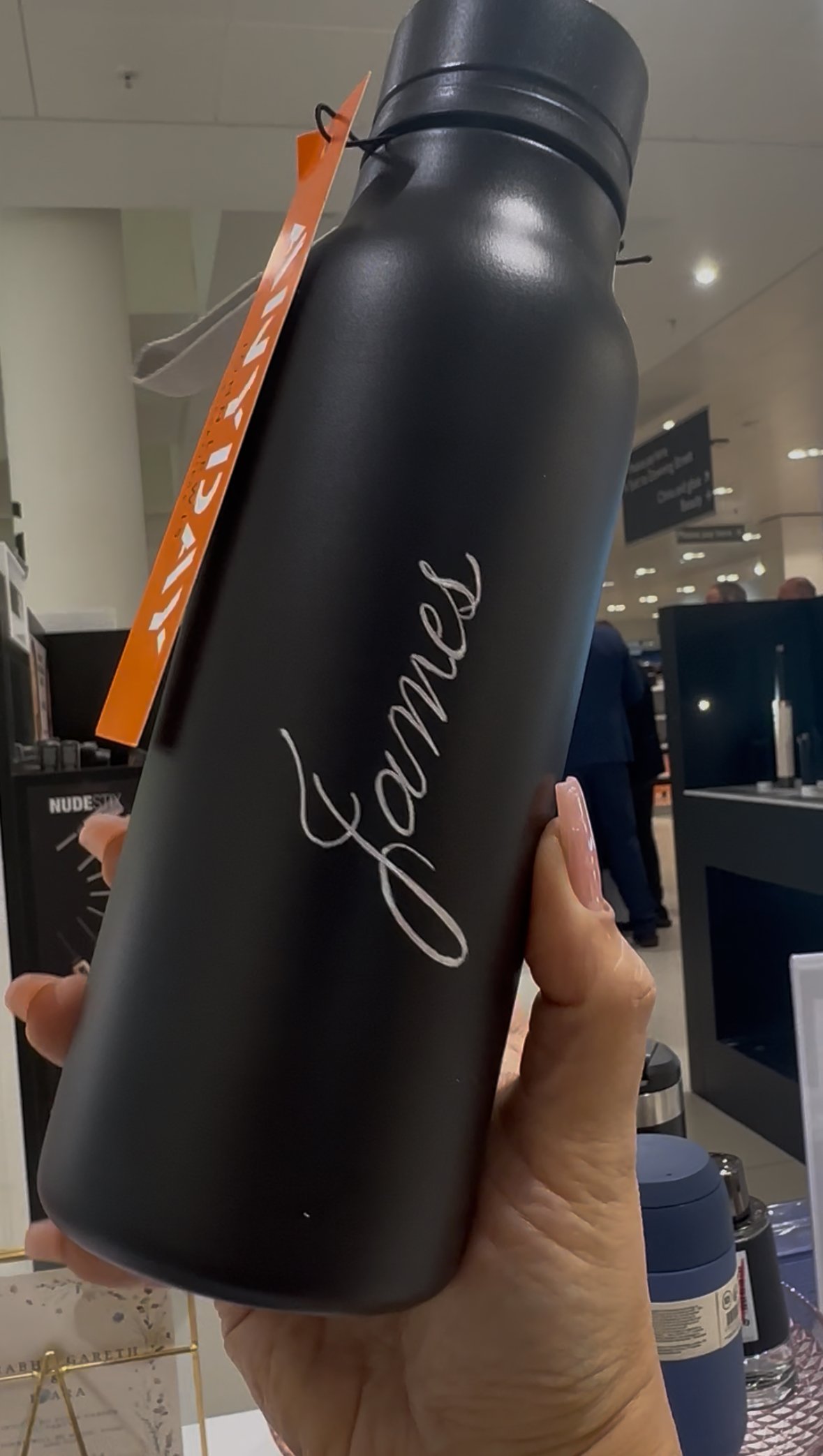 Black water bottle with white cursive 'fame' text held by hand in store aisle.