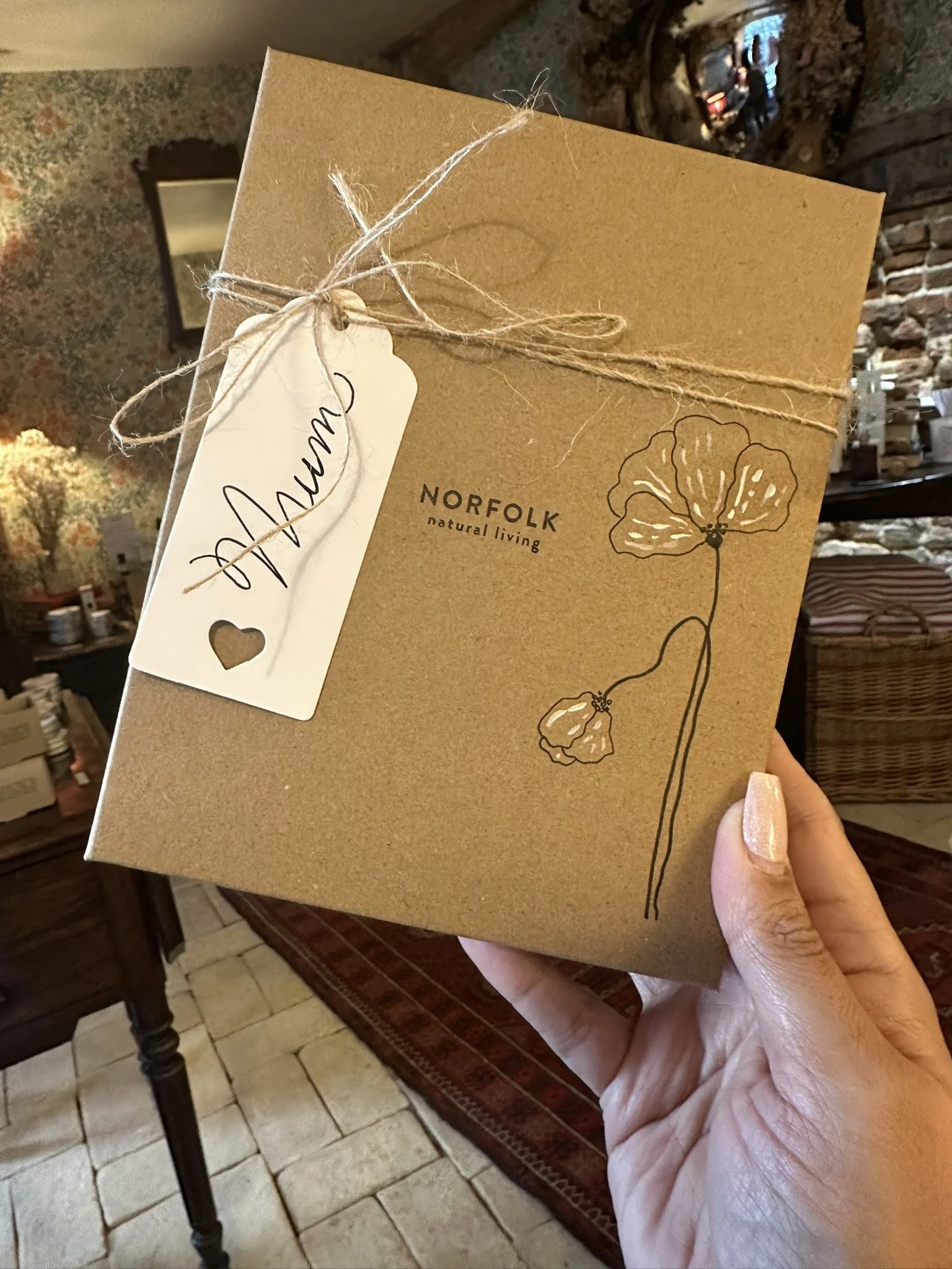 A person holding a cardboard box with a floral design and text reading 'Norfolk natural living.' The box is tied with twine and has a gift tag that says 'Mom' with a small heart cutout. The background shows a cozy interior with a brick fireplace and 