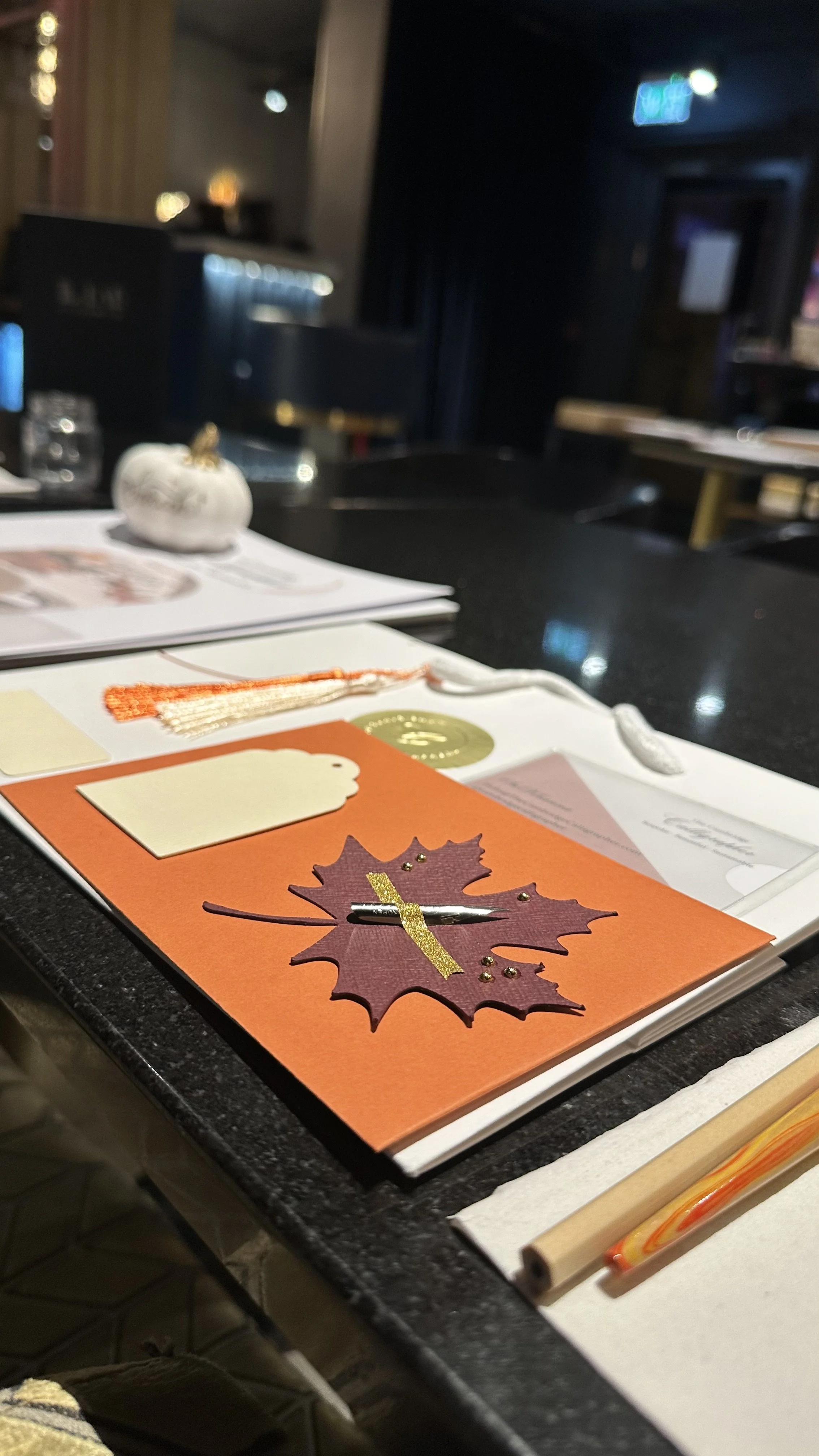 A table with a fall-themed card featuring a maple leaf with glitter and small beads, a white pumpkin, chopsticks, and some papers in a dimly lit restaurant setting.