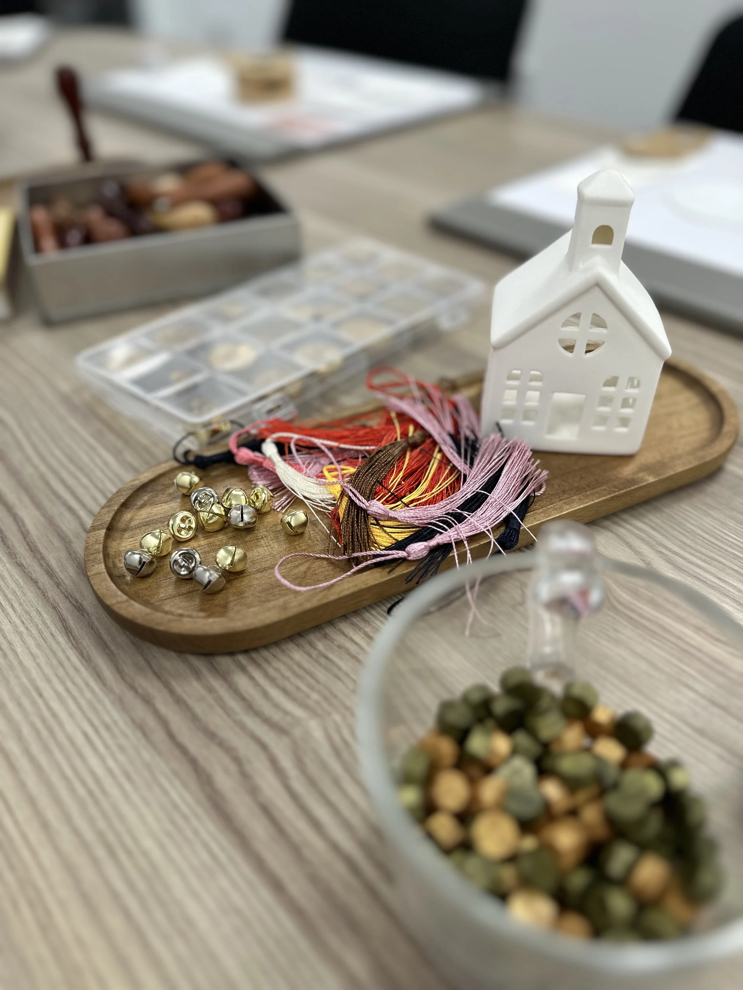 A wooden tray with embroidery threads, small bells, and a white ceramic house decoration, on a wooden table with various craft supplies in the background.