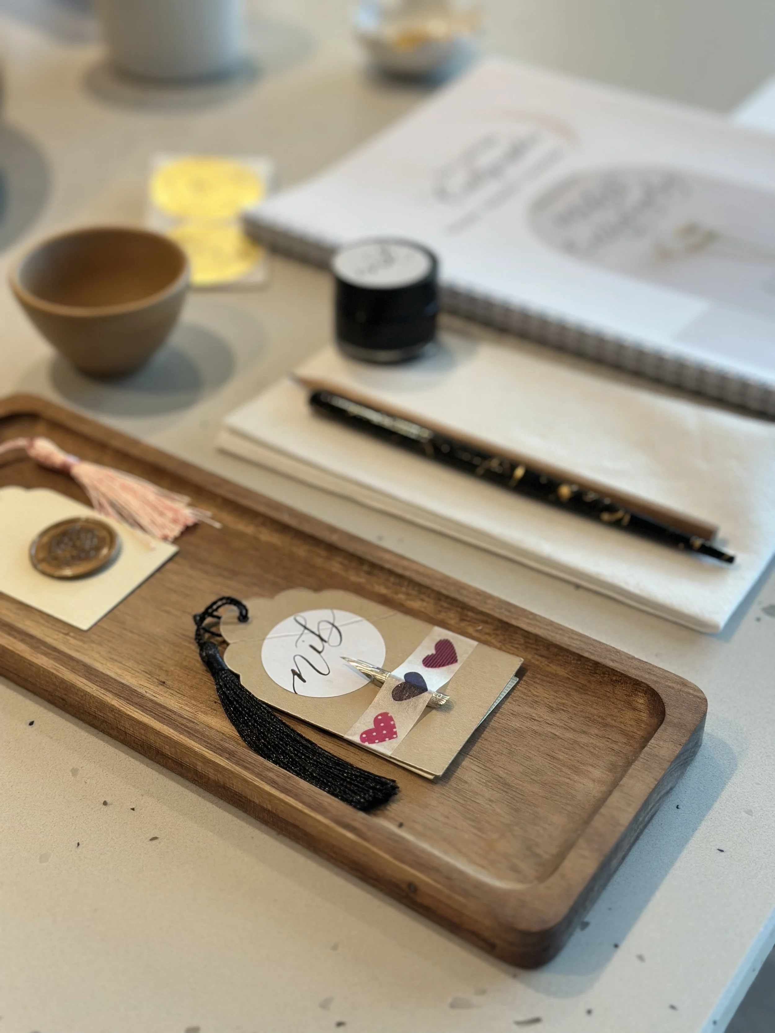 A wooden tray with decorative items including a small black tassel, a handwritten note, and colorful heart stickers, placed on a light-colored surface.