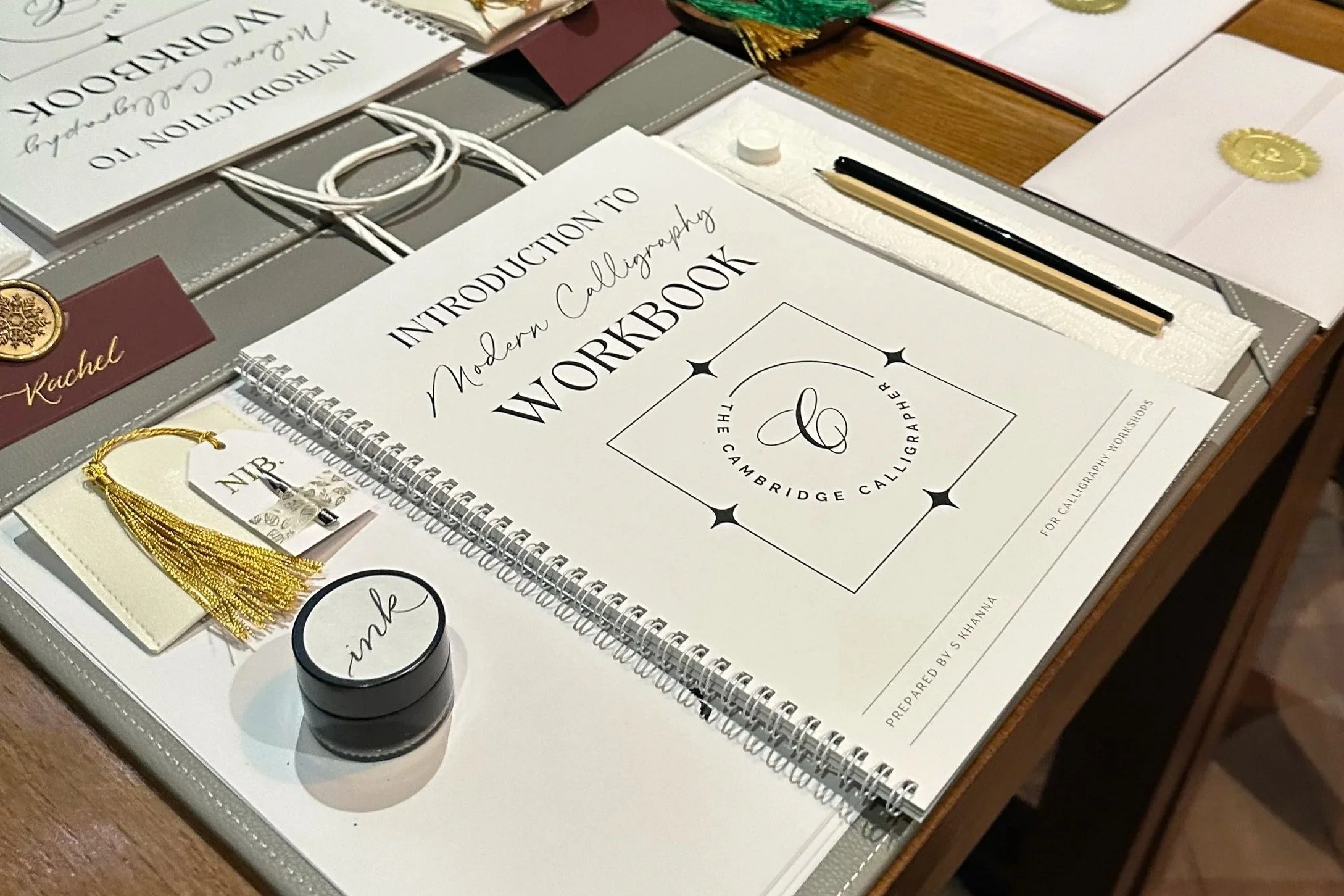 A calligraphy workshop setup with a workbook titled 'Introduction to Modern Calligraphy Workshop,' a pen, a ink pot labeled 'ink,' a small gold tassel, and various calligraphy supplies on a table.