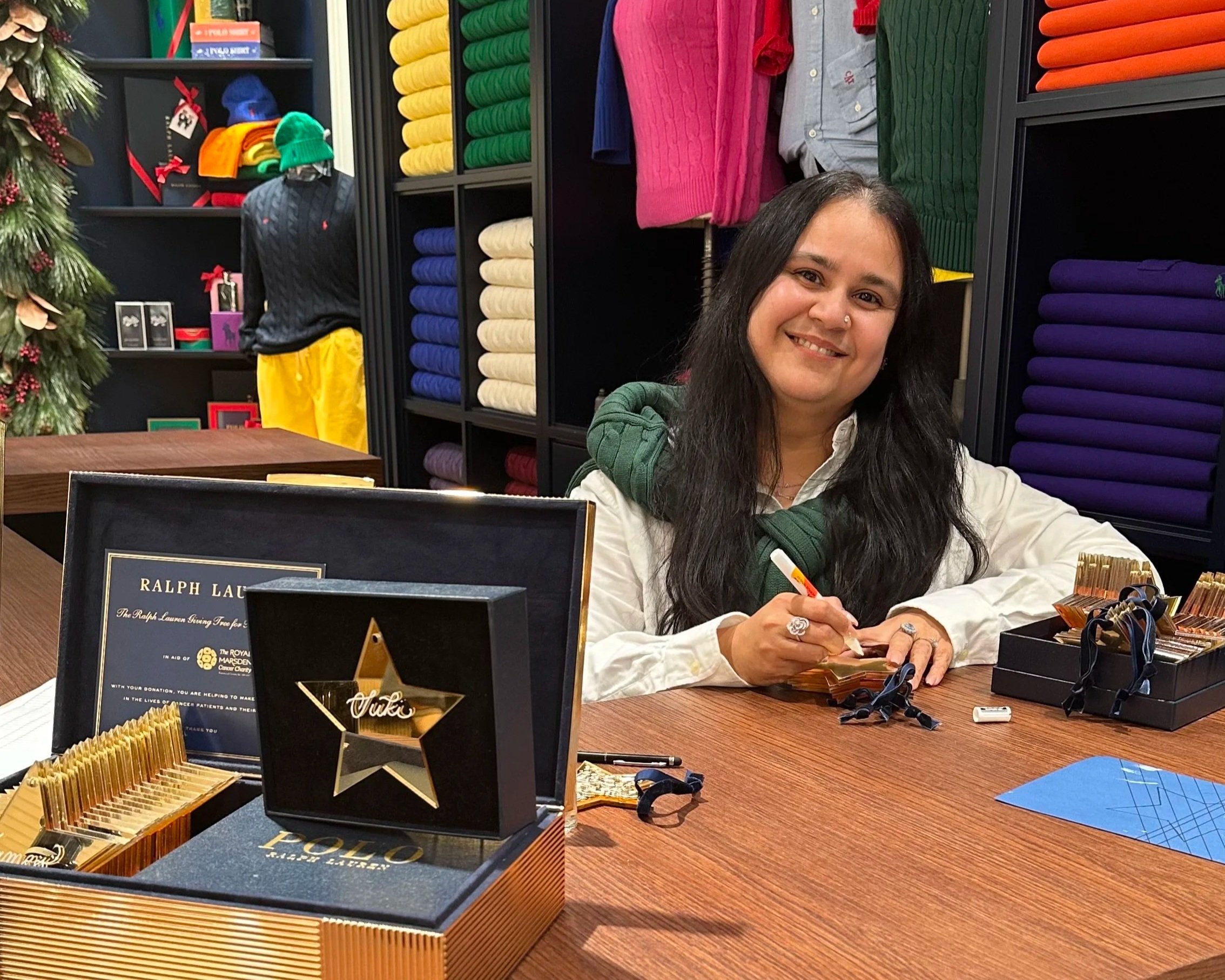 A woman with long black hair wearing a white shirt, sitting at a table signing autographs at a gift shop, with colorful folded sweaters on shelves behind her and a decorated Christmas tree on the left.