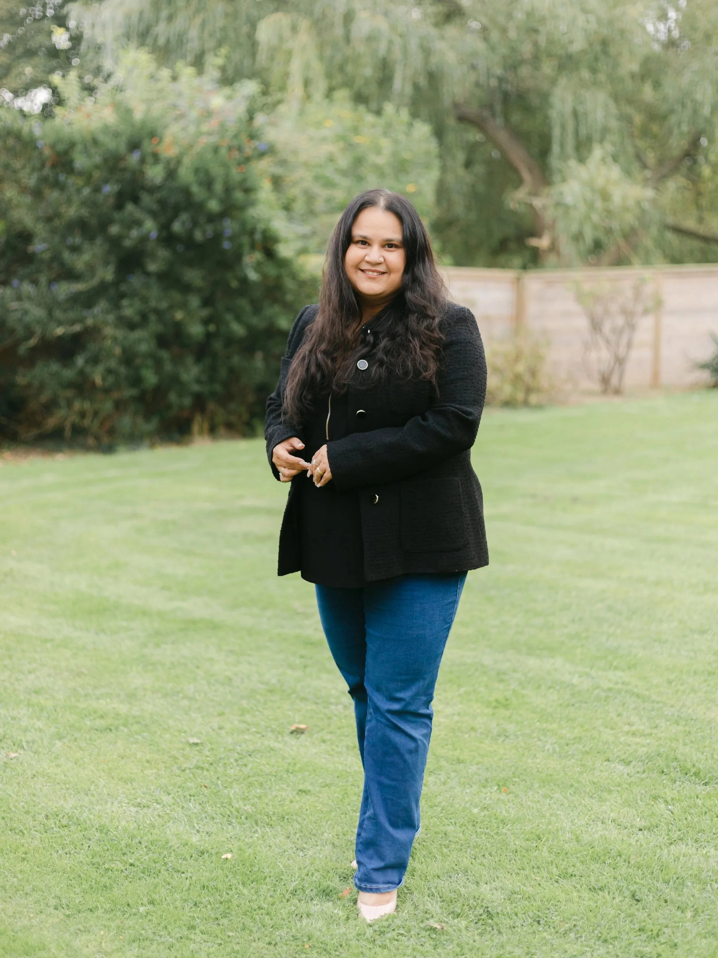 A woman with long dark hair wearing a black blazer and blue jeans standing on a grassy area with trees and a wooden fence in the background.