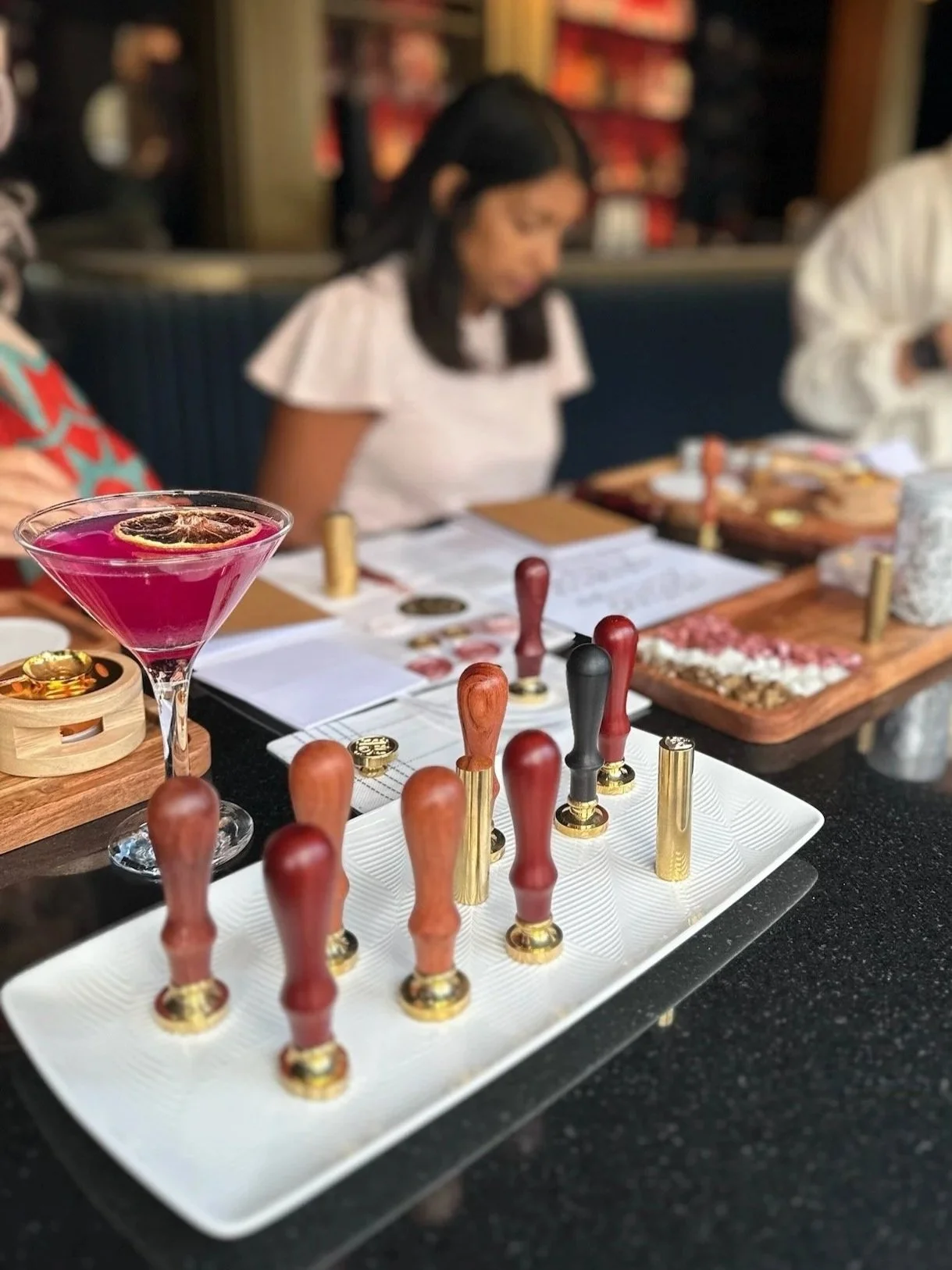 Board game pieces resembling small stamps with wooden handles and gold bases, placed on a white tray, along with a pink cocktail garnished with a lemon slice, on a table set for a game night.