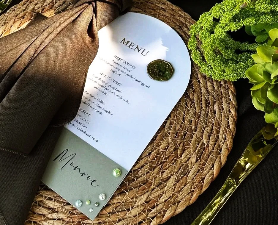 A wedding menu on a white card with a wax seal, placed on a woven placemat next to a chocolate brown napkin, green flowers, and gold cutlery.