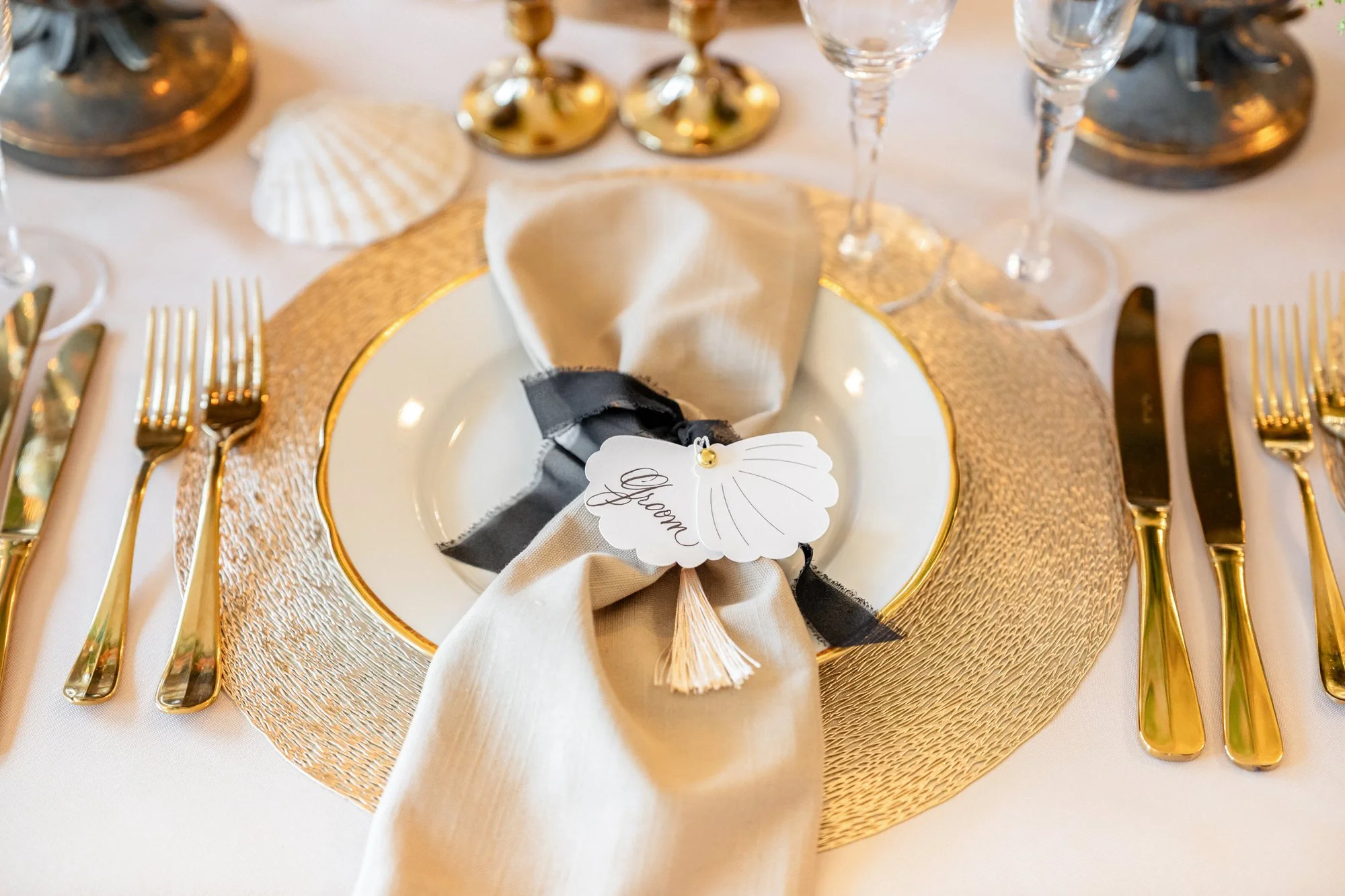 Elegant table setting with gold cutlery, glassware, a scalloped name tag that says "Groom," and seashell decorations.