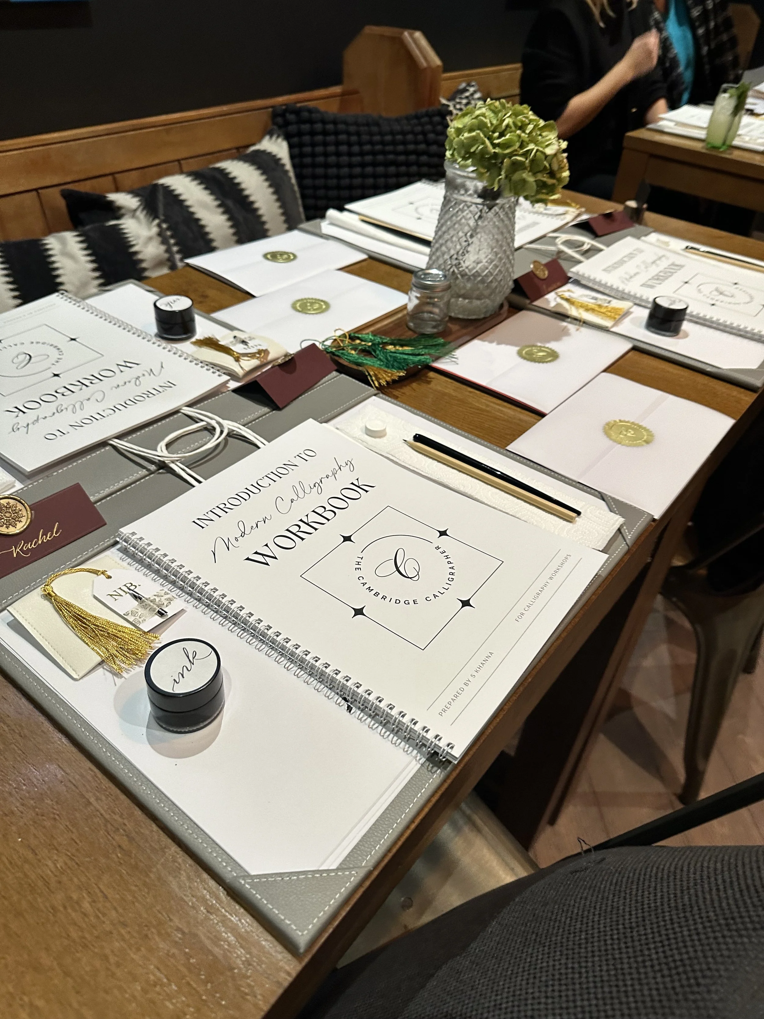 Table set for calligraphy workshop with notebooks, calligraphy supplies, and a flower vase.