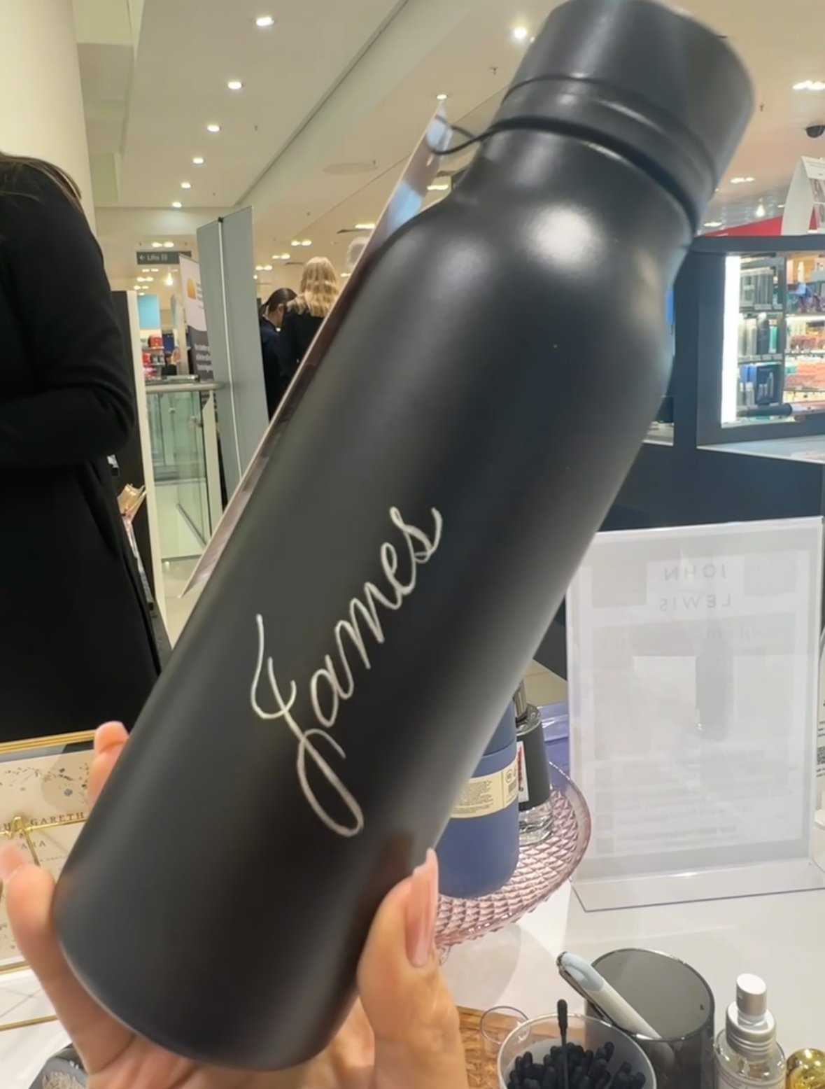 Black travel mug with white cursive writing that says 'fame' being held in a retail store with people and shelves in the background.