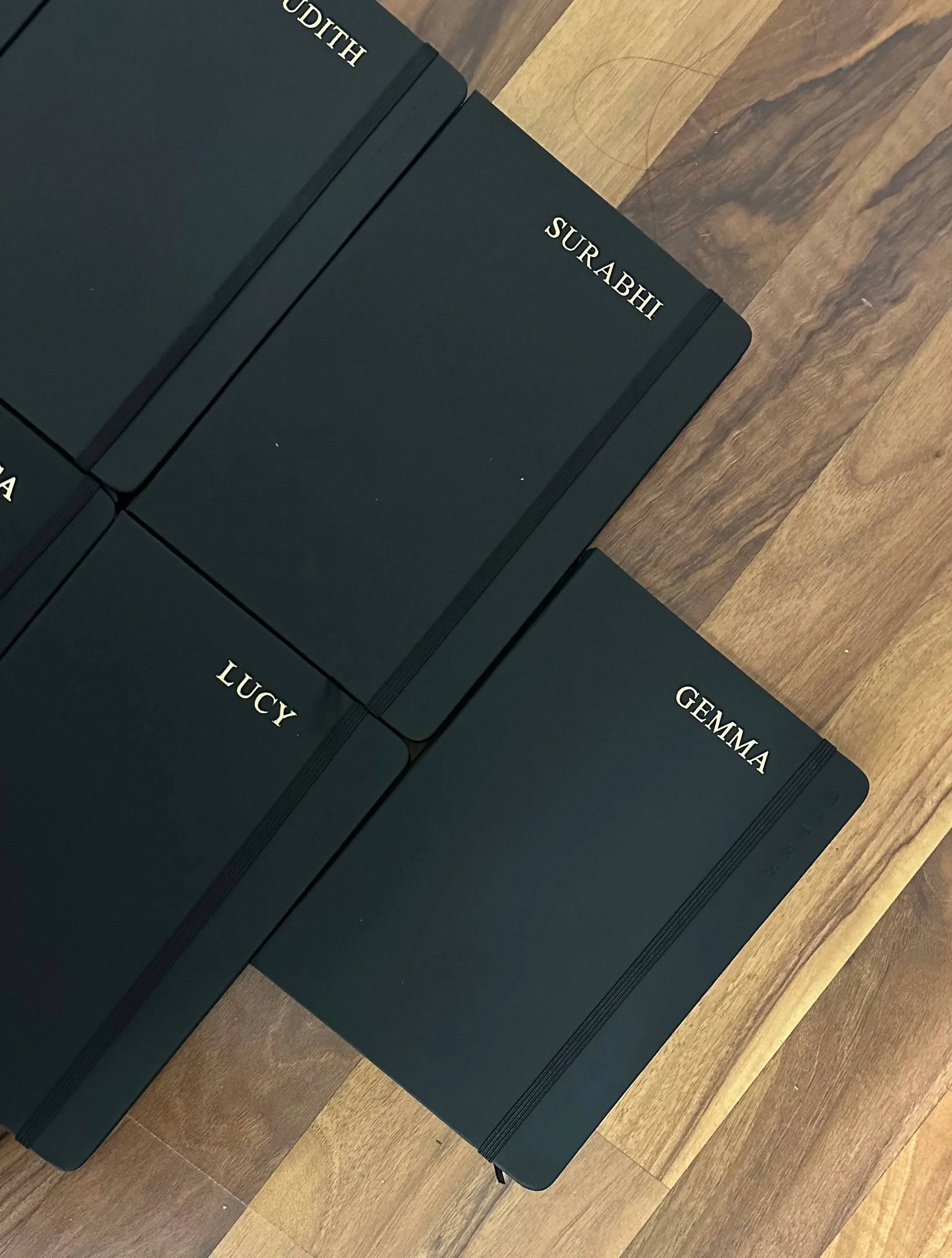 Four closed black notebooks with white embossed names 'GEMMA', 'LUCY', 'SURAHI', and 'KHADIJAH' arranged on a wooden surface.