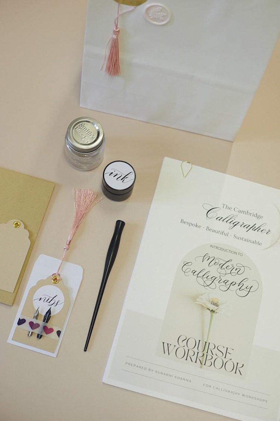 A calligraphy course workbook titled 'Modern Calligraphy' on a table with calligraphy pens, a jar of ink, a charm with the word 'nib', and a paper bag with a pink tassel and seal.