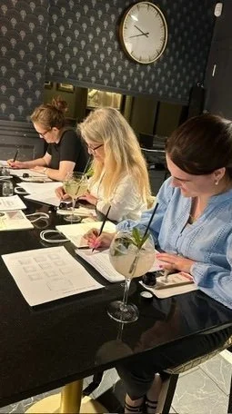 Three women sitting at a table, taking notes, with drinks in front of them, in a room with a patterned wall and a clock on the wall.