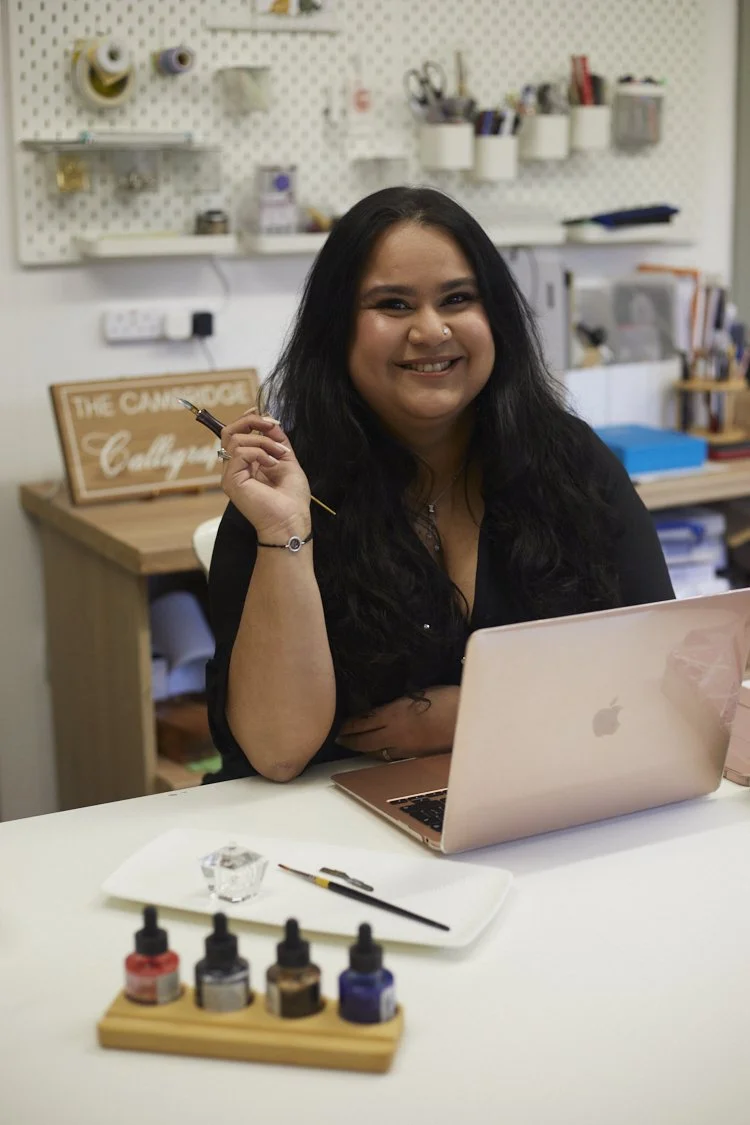 Cambridge Calligrapher (Su) sitting at her desk behind a laptop, holding a calligraphy pen