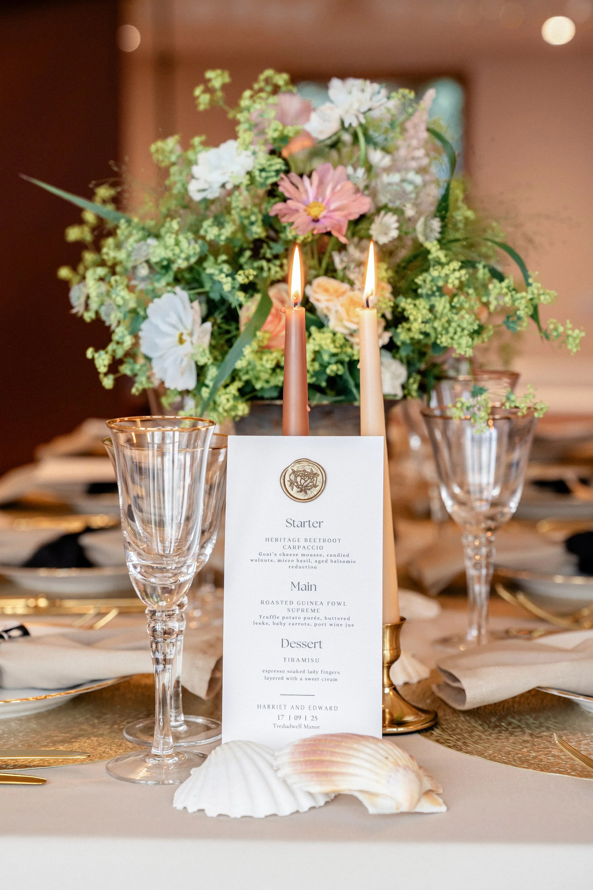 Elegant table setting with a floral centerpiece, two lit candles, clear glassware, a printed menu card, seashells, and gold cutlery.