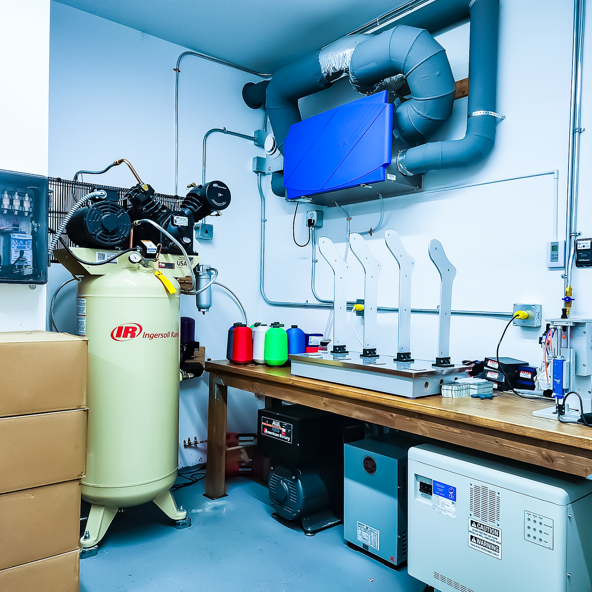 A machine room containing a yellow air compressor, several spools of colored thread or string, electrical equipment, boxes, and various mechanical and electronic devices on a wooden workbench. The room has blue piping and ventilation system on the wa