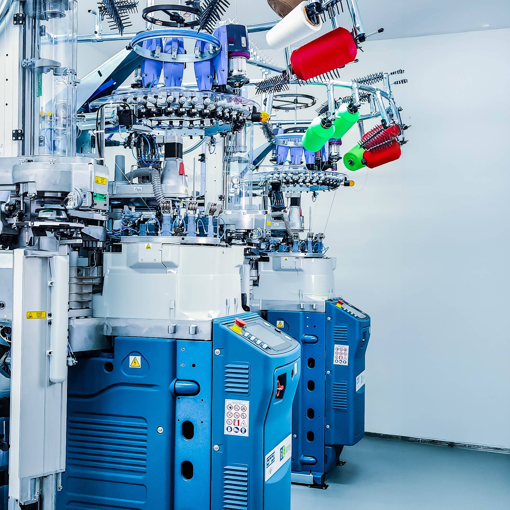 Close-up of a complex industrial machinery system with various metallic components, wiring, and colorful spools of sewing thread or wire in a clean, well-lit environment.