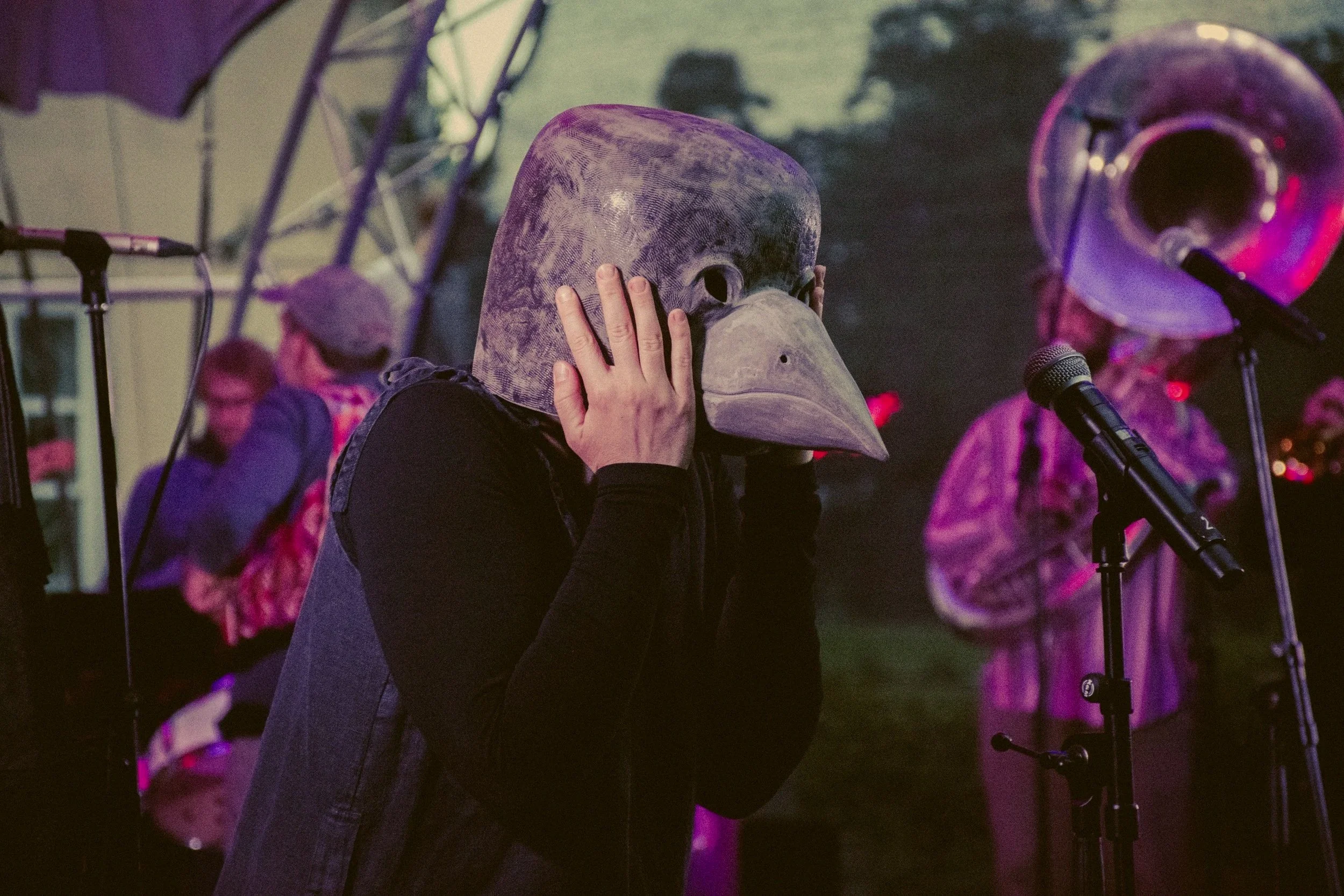 Wearable performance art: Monika Dombrauskytė in the Bird Mask stands at a microphone with an improvisational orchestra behind them.