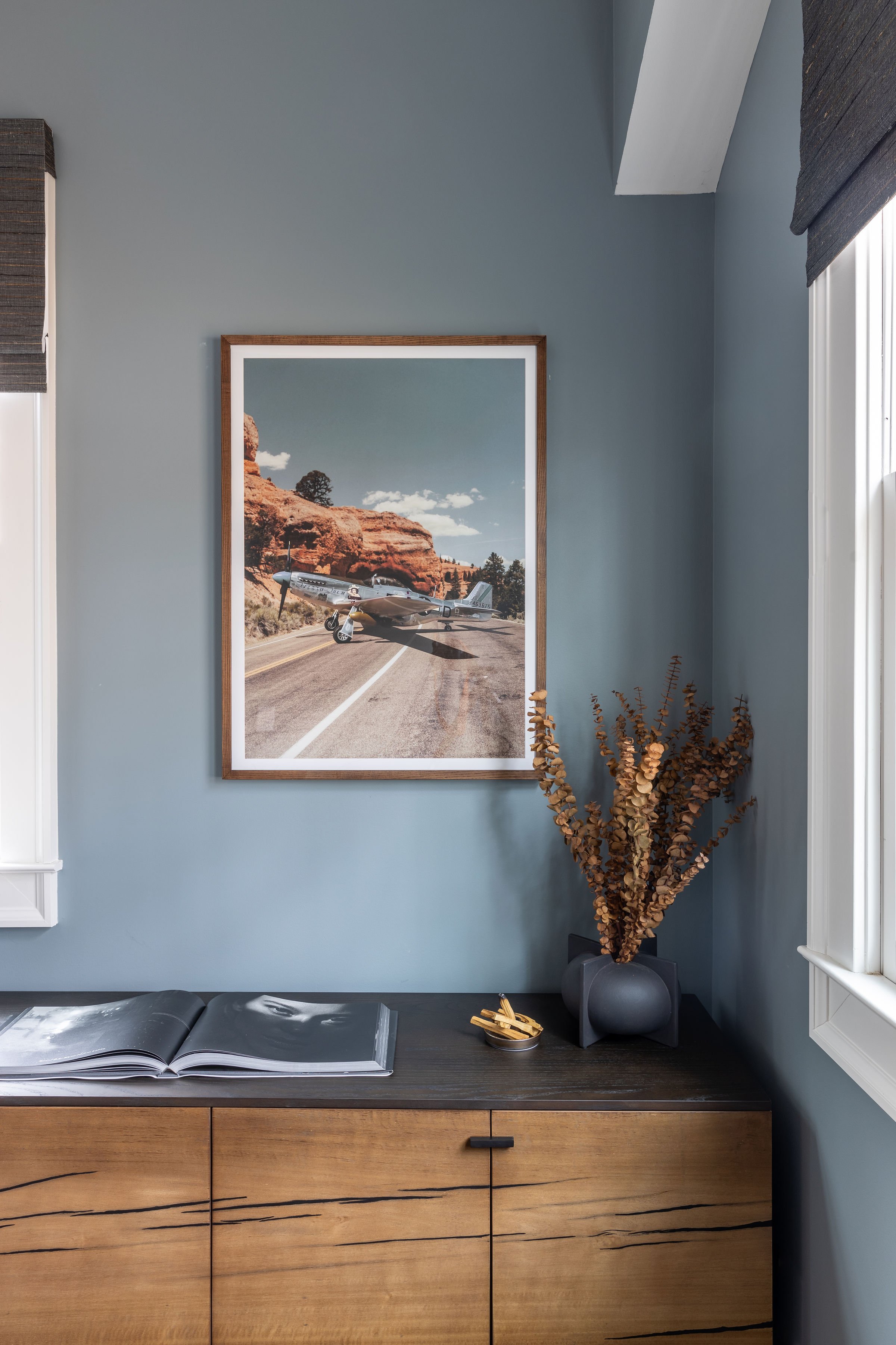 modern office design boosting soft earthly blue walls with a masculine dark wood credenza. On display are a photograph of an airplane, a vase with dried eucalyptus leaves, santal wood and an open photography book.