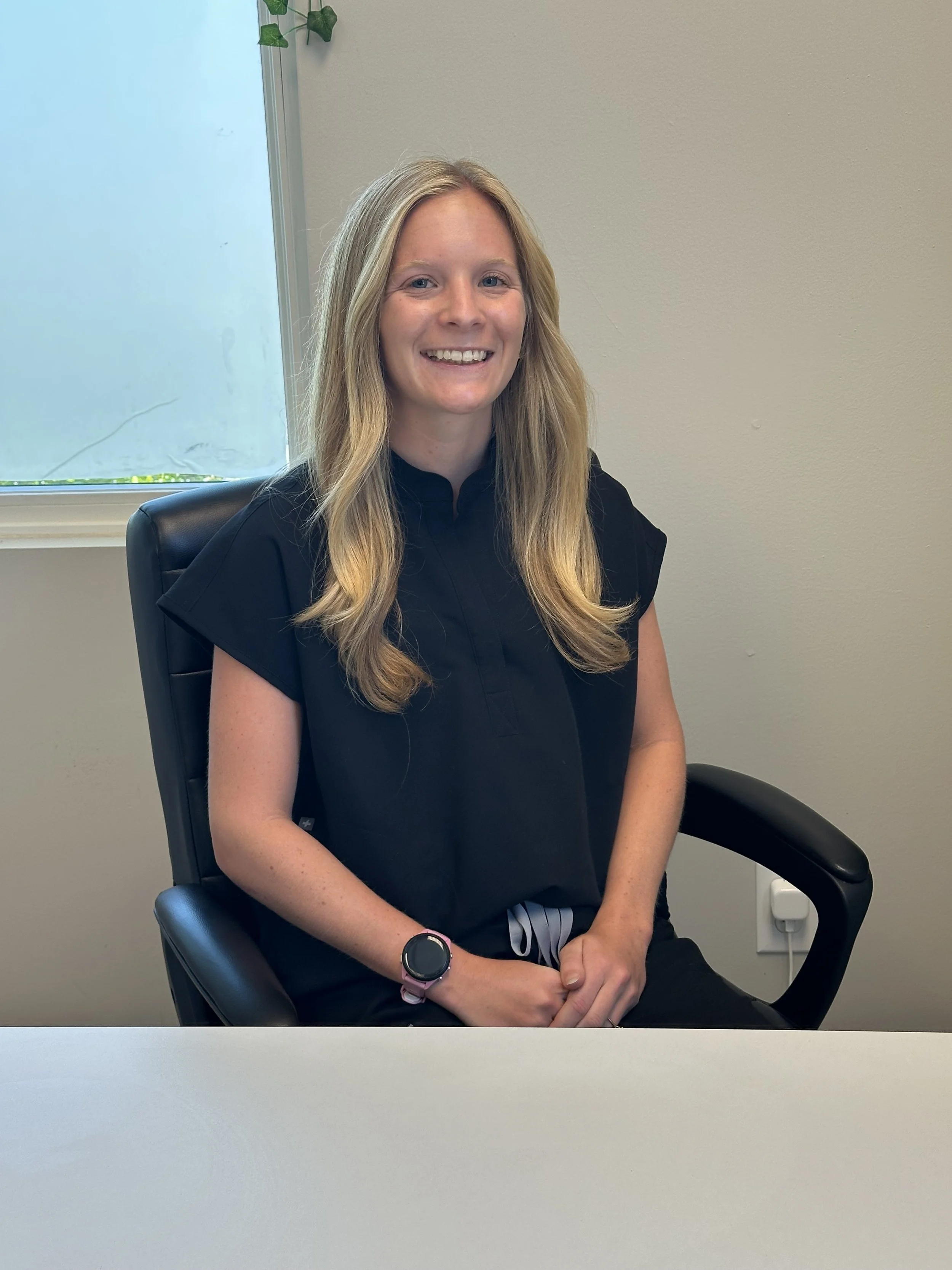 A woman with long blonde hair smiling while sitting in a black office chair in front of a white desk and beige wall with a window and green plant.