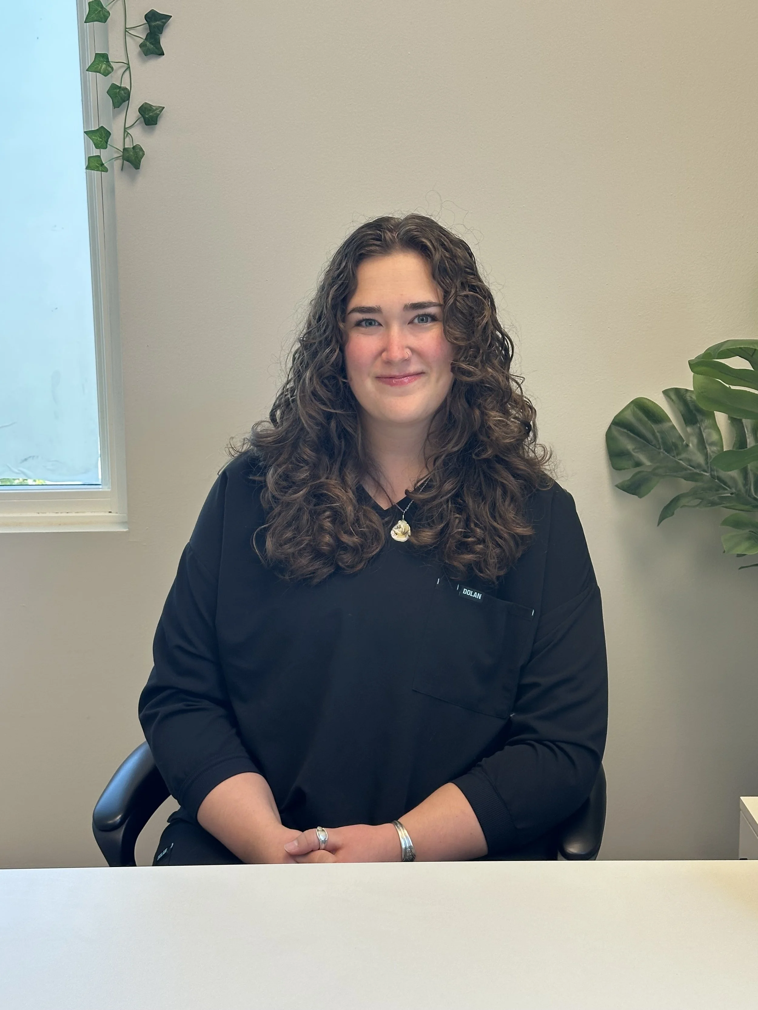 A woman with curly brown hair and blue eyes sitting at a desk, wearing a black jacket, silver jewelry, and smiling, with a window and a green plant in the background.