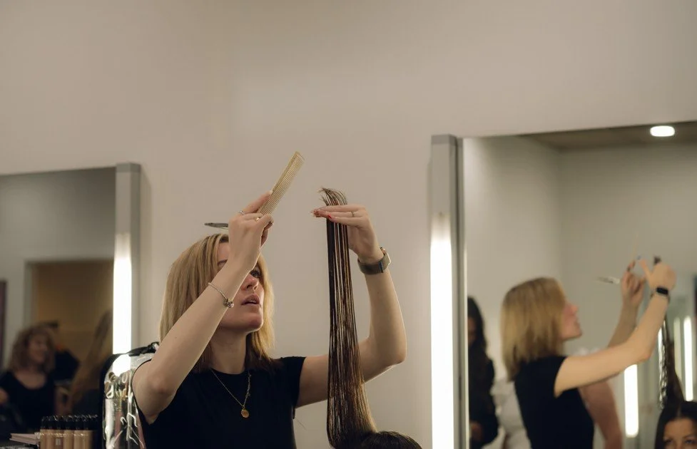 A hairstylist holding hair extensions and a comb while working in a salon, with a mirror reflecting her and her client.