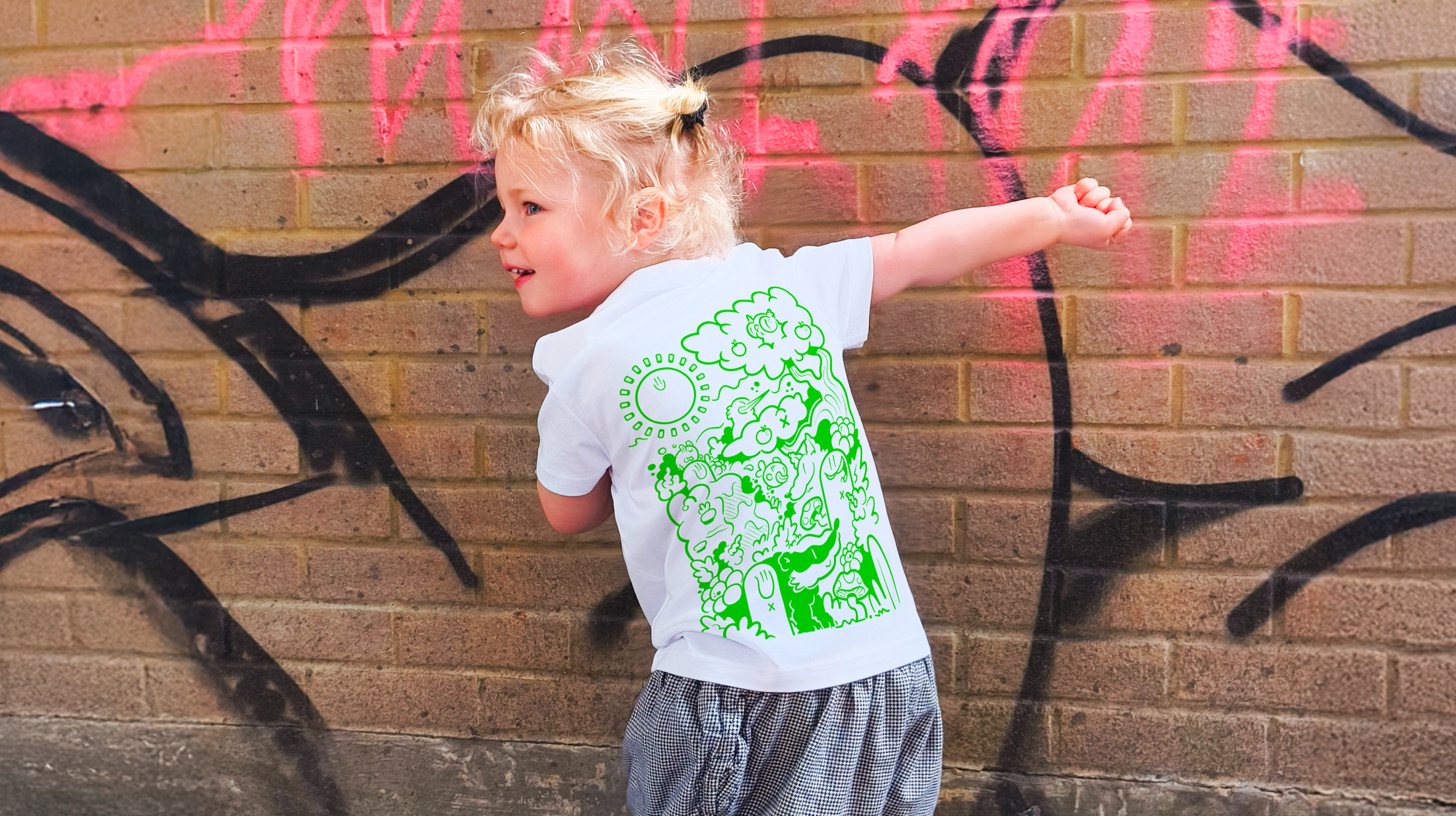 A young child with blonde curly hair and wearing a white T-shirt with green graphic design, standing against a brick wall with black and pink graffiti, smiling and extending their right arm.