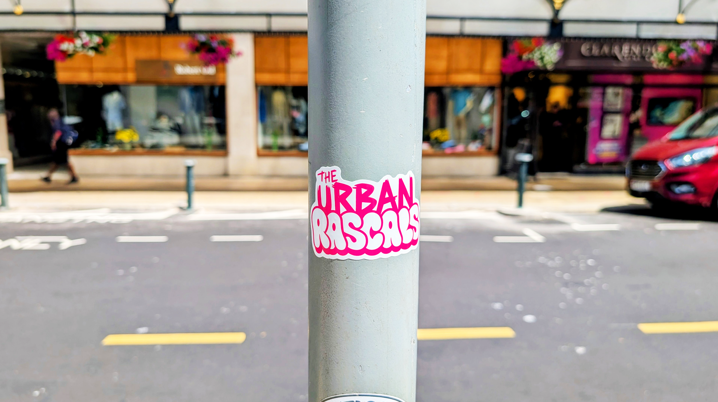 Pink and white sticker on a gray pole that says "The Urban Rascals" in stylized font.