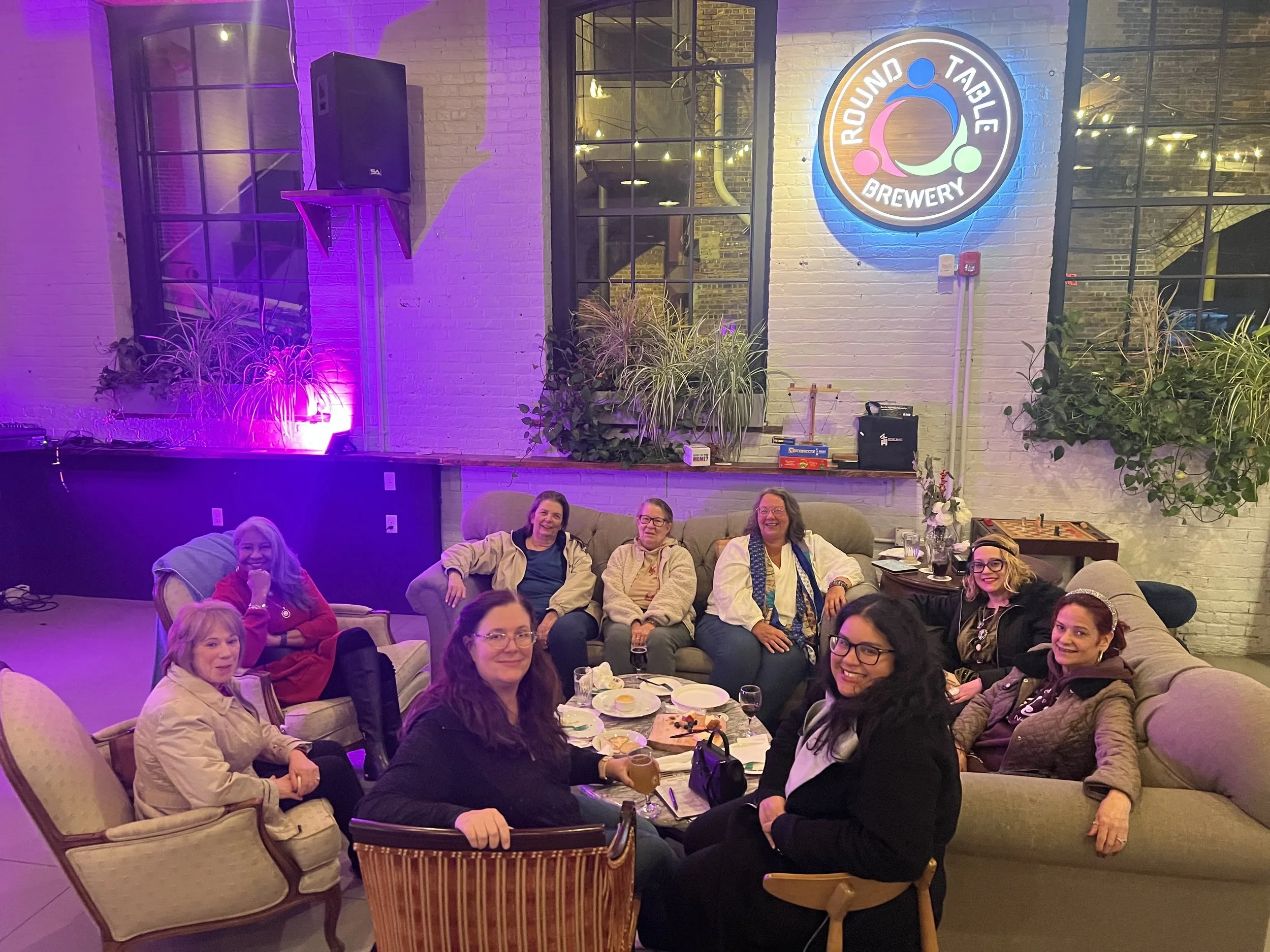 A group of women sitting on sofas and chairs at a social gathering in a brewery with a neon sign reading 'Round Table Brewery' on the wall.