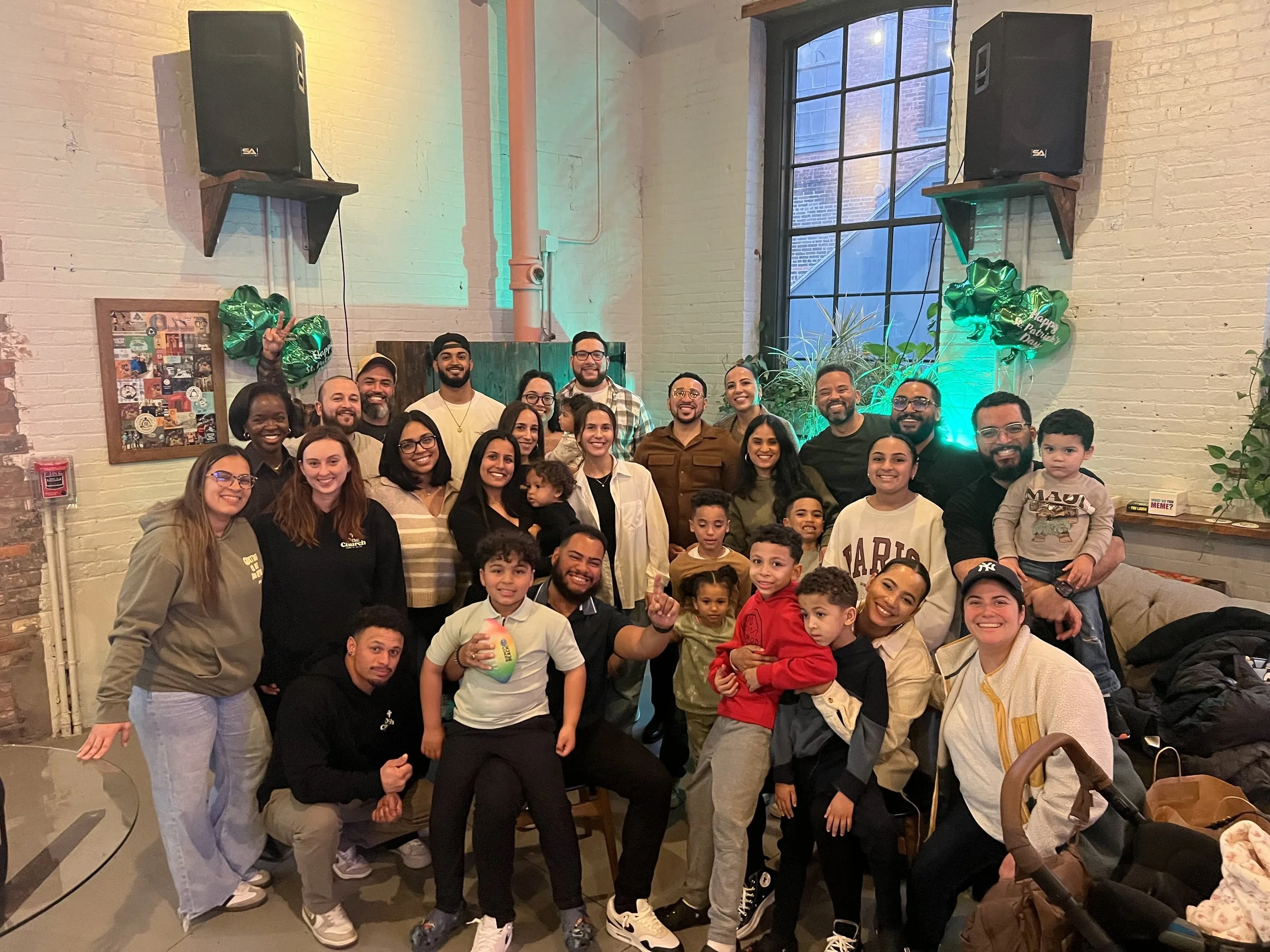 Group photo of diverse adults and children celebrating St. Patrick's Day indoors with green balloons, plants, and colorful lighting.
