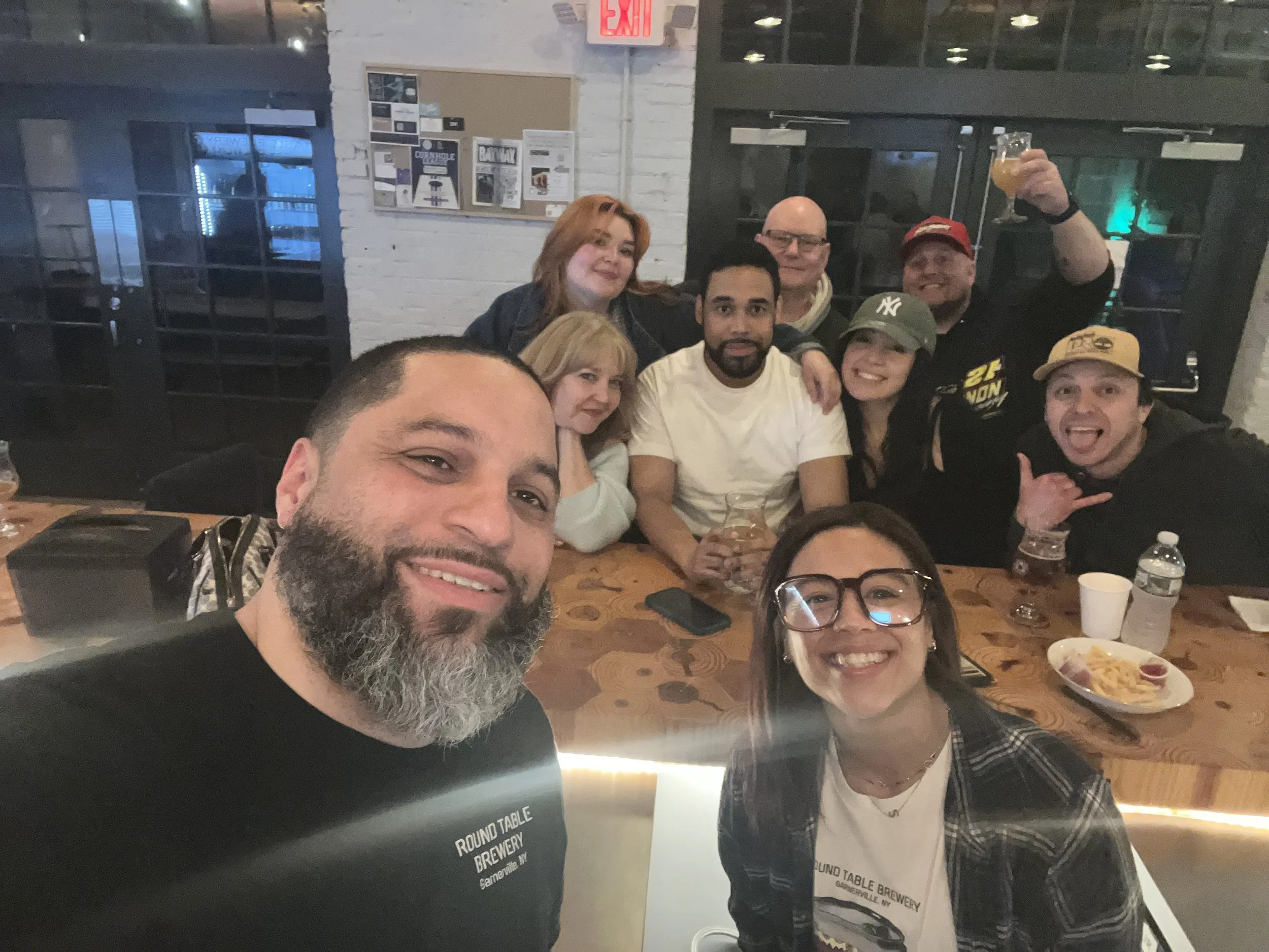 Group of ten friends celebrating at a bar or brewery, sitting around a wooden table with drinks and food, smiling and taking a selfie. The setting has a modern, industrial style with brick walls and black-framed doors.
