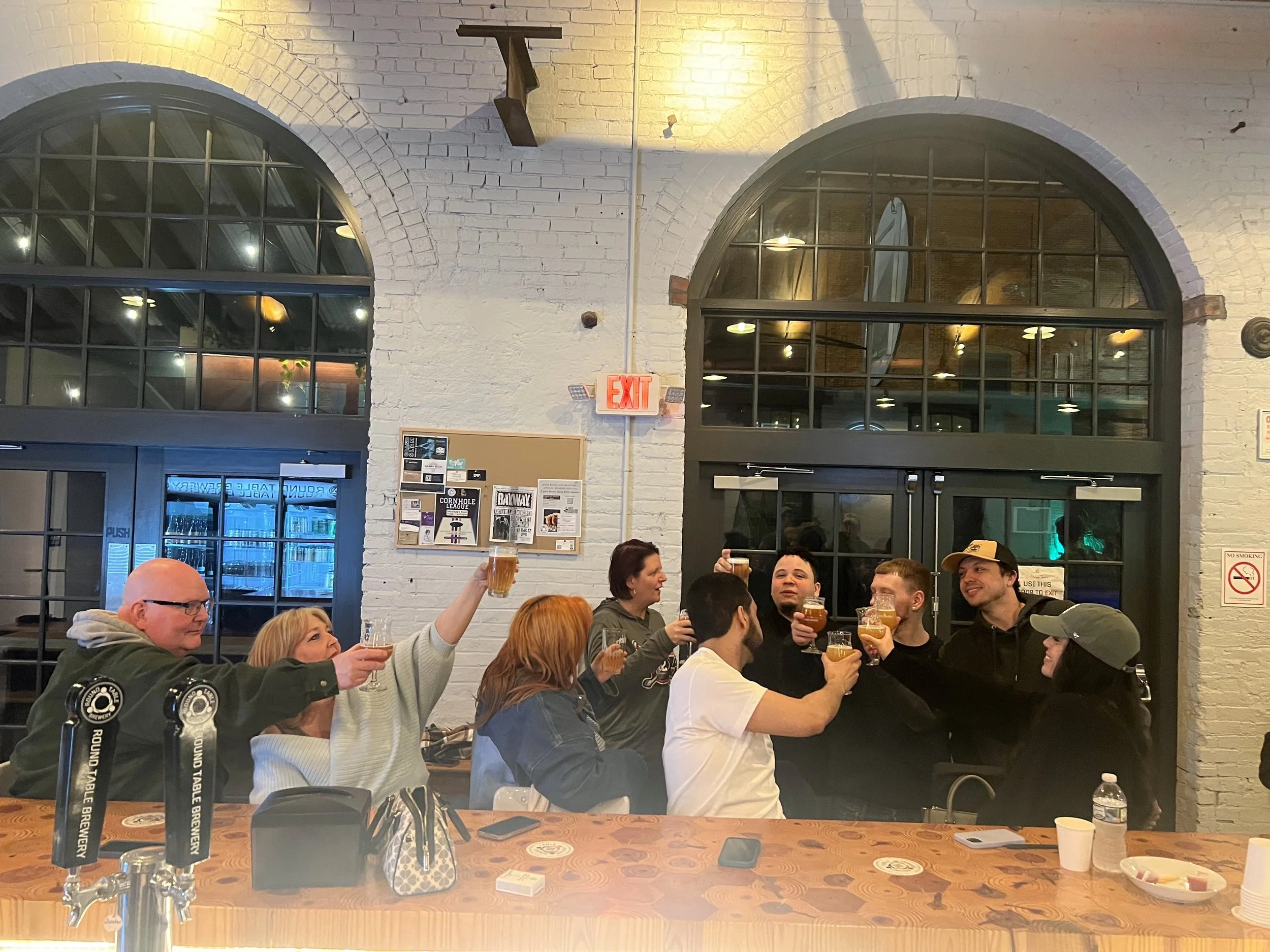 A group of people gathered inside a brewery or bar, raising their glasses for a toast. The setting has brick walls, large arched windows, and a long wooden counter with taps and items on it.