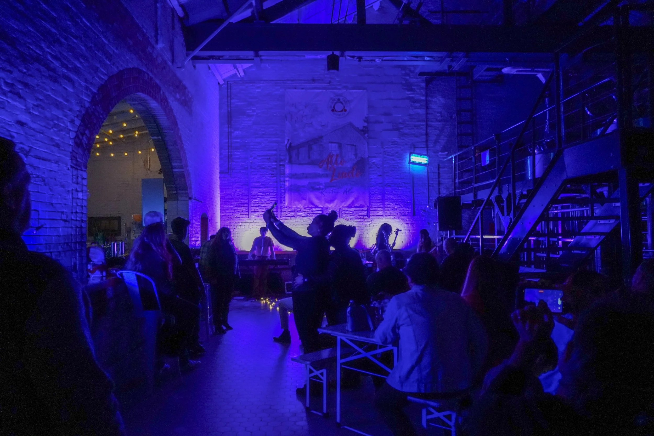 People gathered in a dimly lit venue with purple and blue lighting, watching a live music performance on stage with a brick wall and a large banner behind them. Some individuals are sitting, others are standing or dancing, with one person raising their arm. There are stairs on the right side and a doorway on the left with string lights.