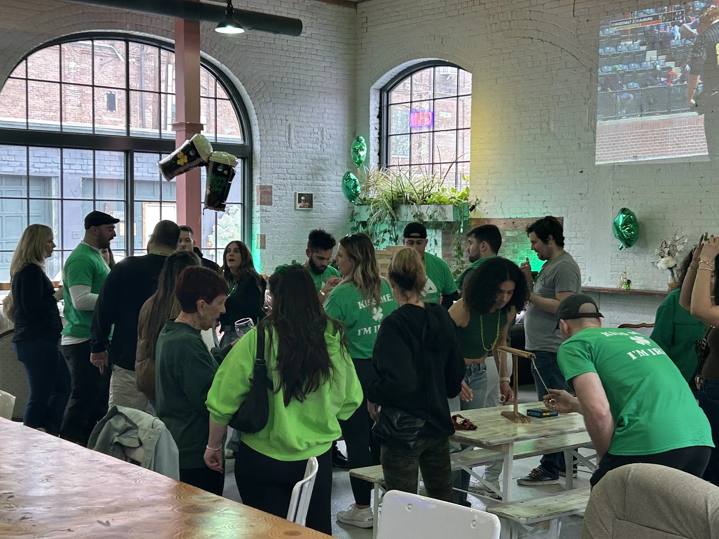 People celebrating St. Patrick's Day in a decorated indoor space, wearing green shirts and hats, with some balloons and a large projected sports game on the wall.
