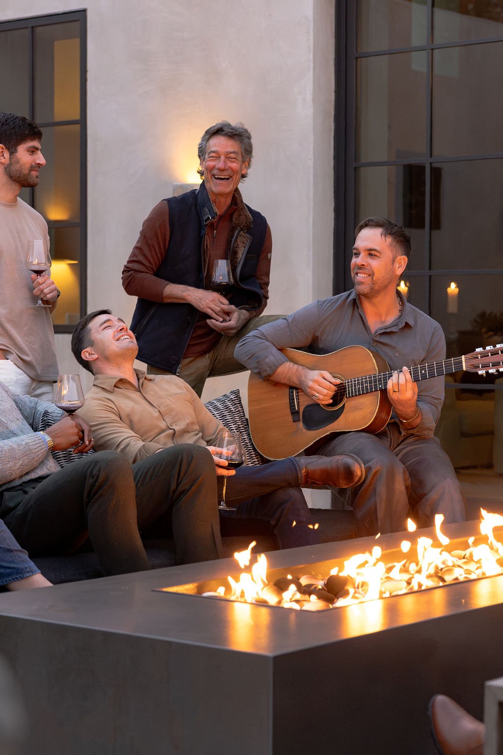 Group of friends enjoying outdoor gathering with guitar, fire pit, and drinks, laughing and chatting.