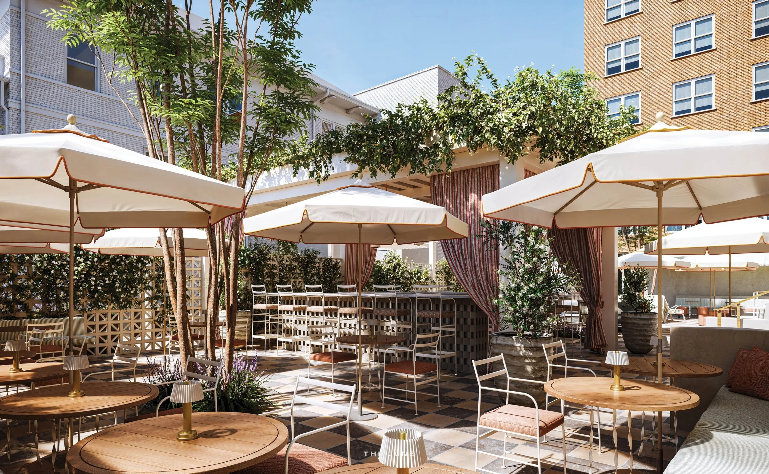 An outdoor patio area with white tables, chairs, large umbrellas, and some trees and plants, surrounded by multi-story buildings.