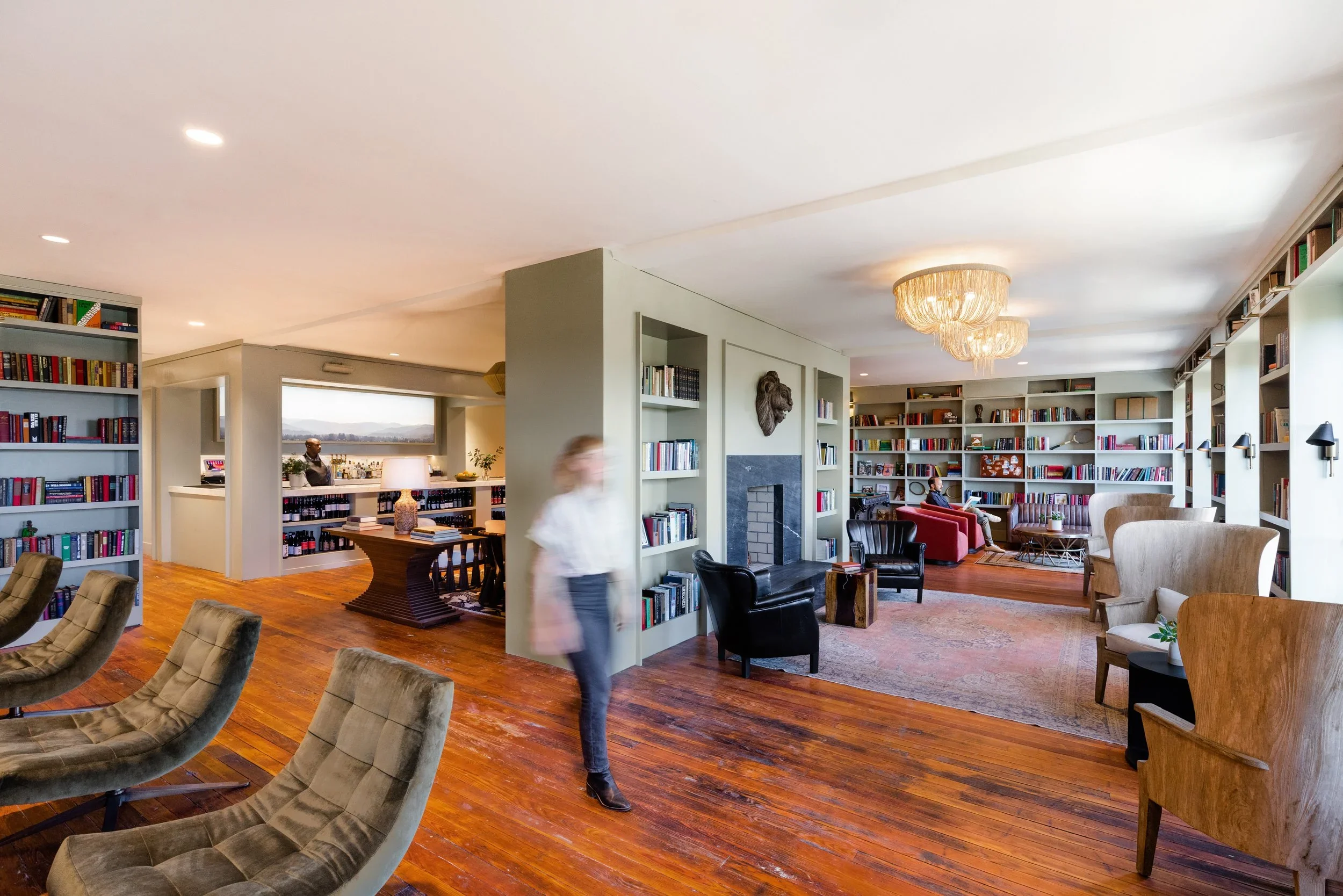 A spacious, well-lit library room with wooden floors, multiple bookshelves filled with books, a seating area with armchairs and a sofa, a fireplace, and chandeliers hanging from the ceiling. There are people walking and sitting in the room.