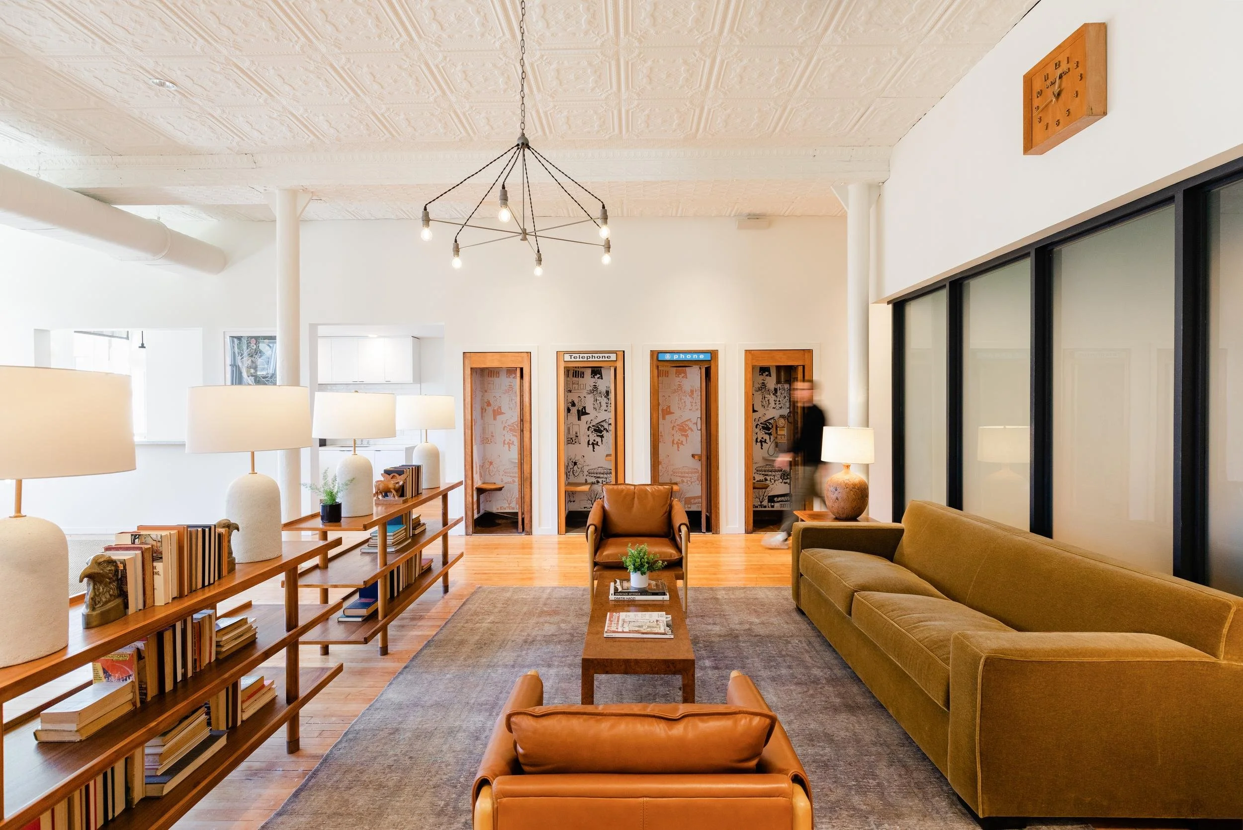 A cozy living room with leather and fabric sofas, wooden coffee table, bookshelf, framed artwork, and a wall clock, with large glass partitions and a modern light fixture.