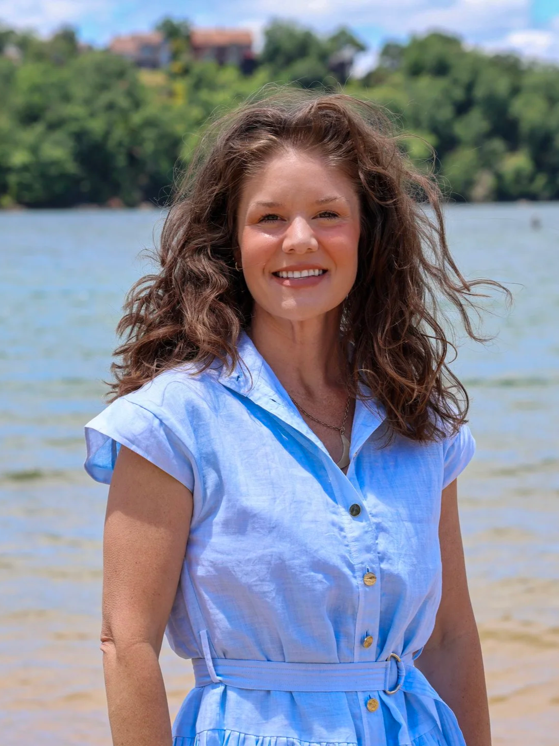 A woman with brown curly hair smiling at the camera outdoors, wearing a light blue button-up dress with a tied waist, standing by a body of water with greenery and houses in the background.