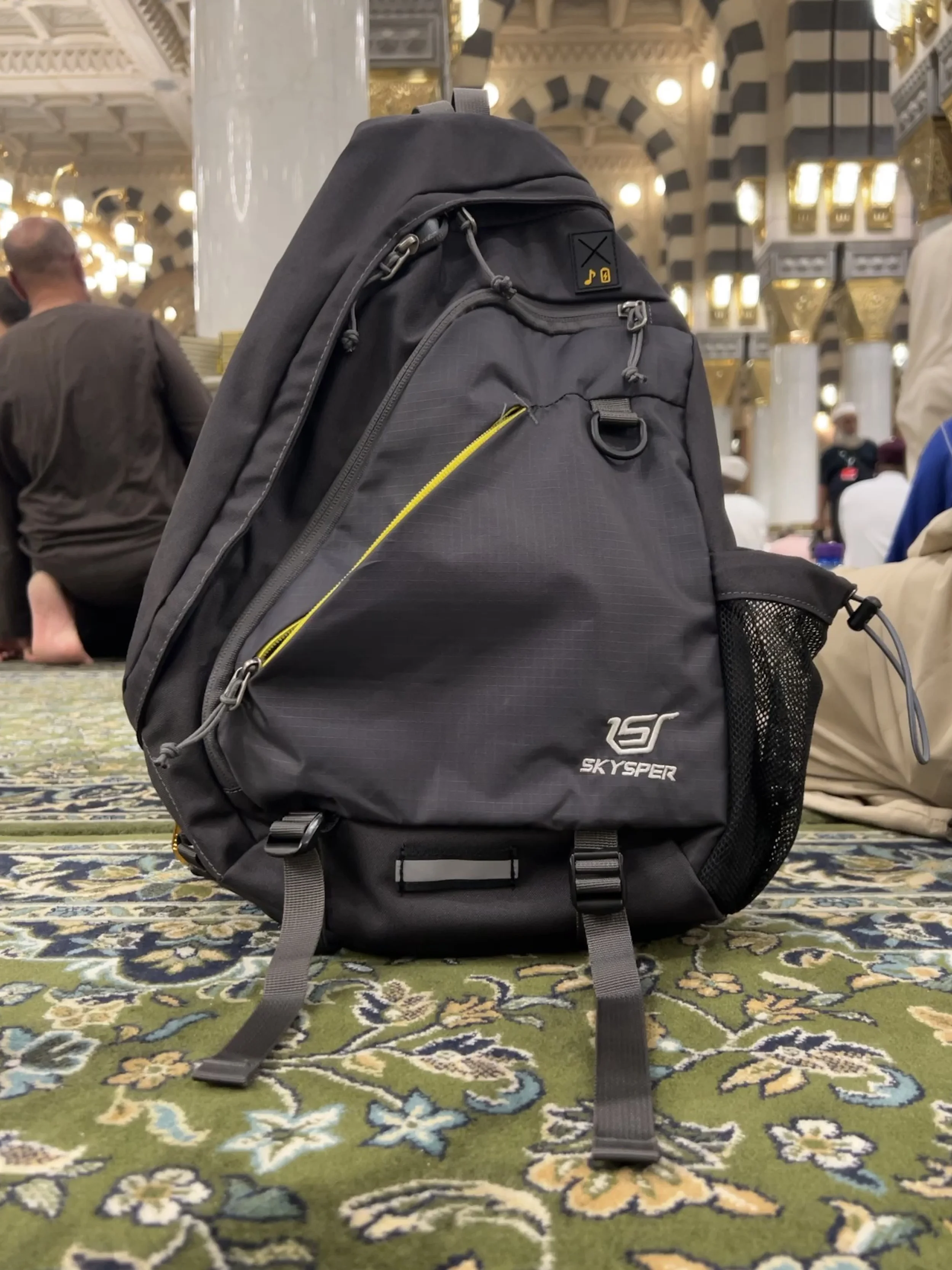 A black Skyser backpack resting on a patterned green carpet in an ornate, well-lit room, with people sitting nearby.