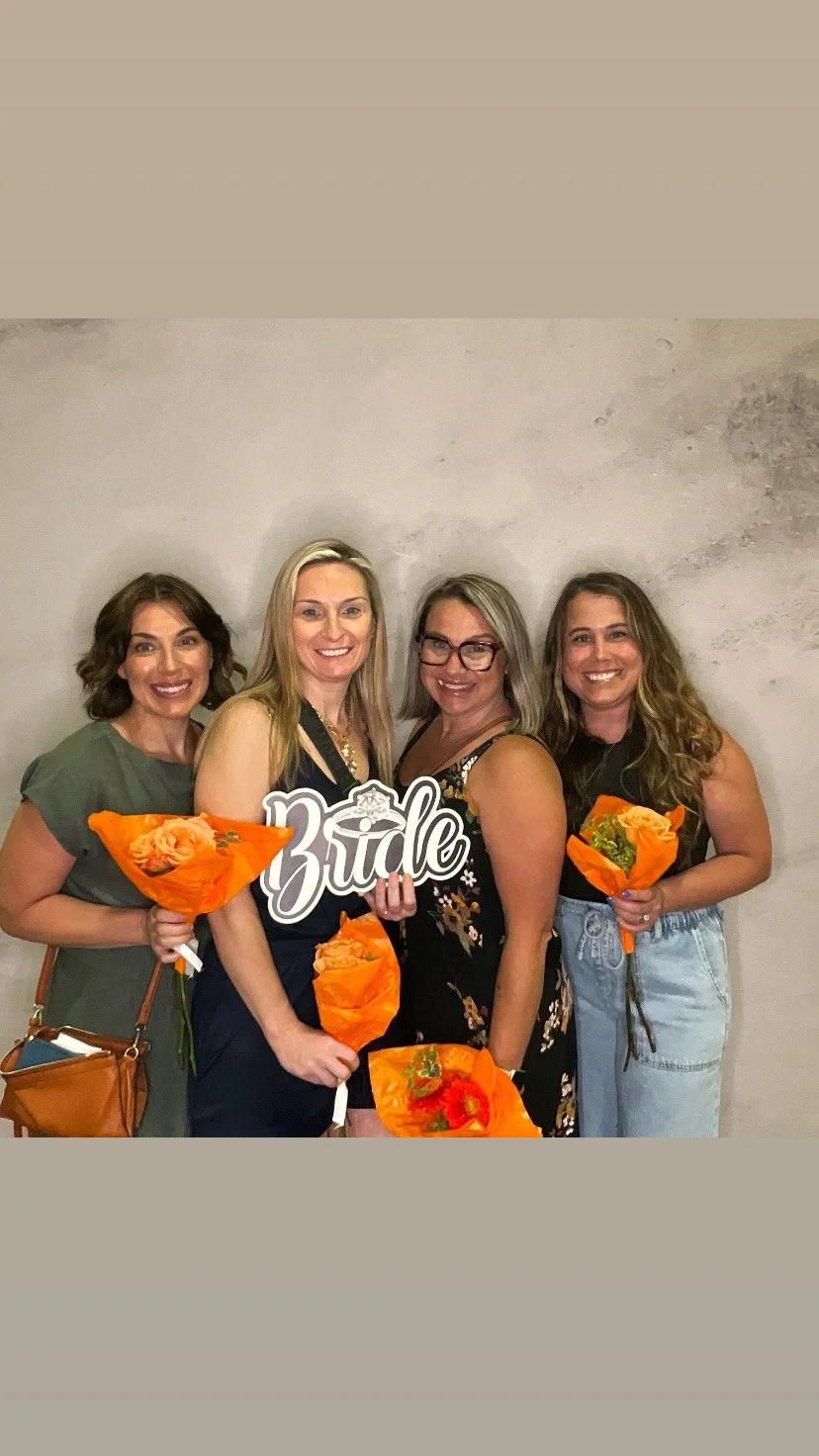 Four women smiling and holding orange bouquets of flowers, standing side by side against a plain wall. One woman is holding a sign that reads 'bride.'