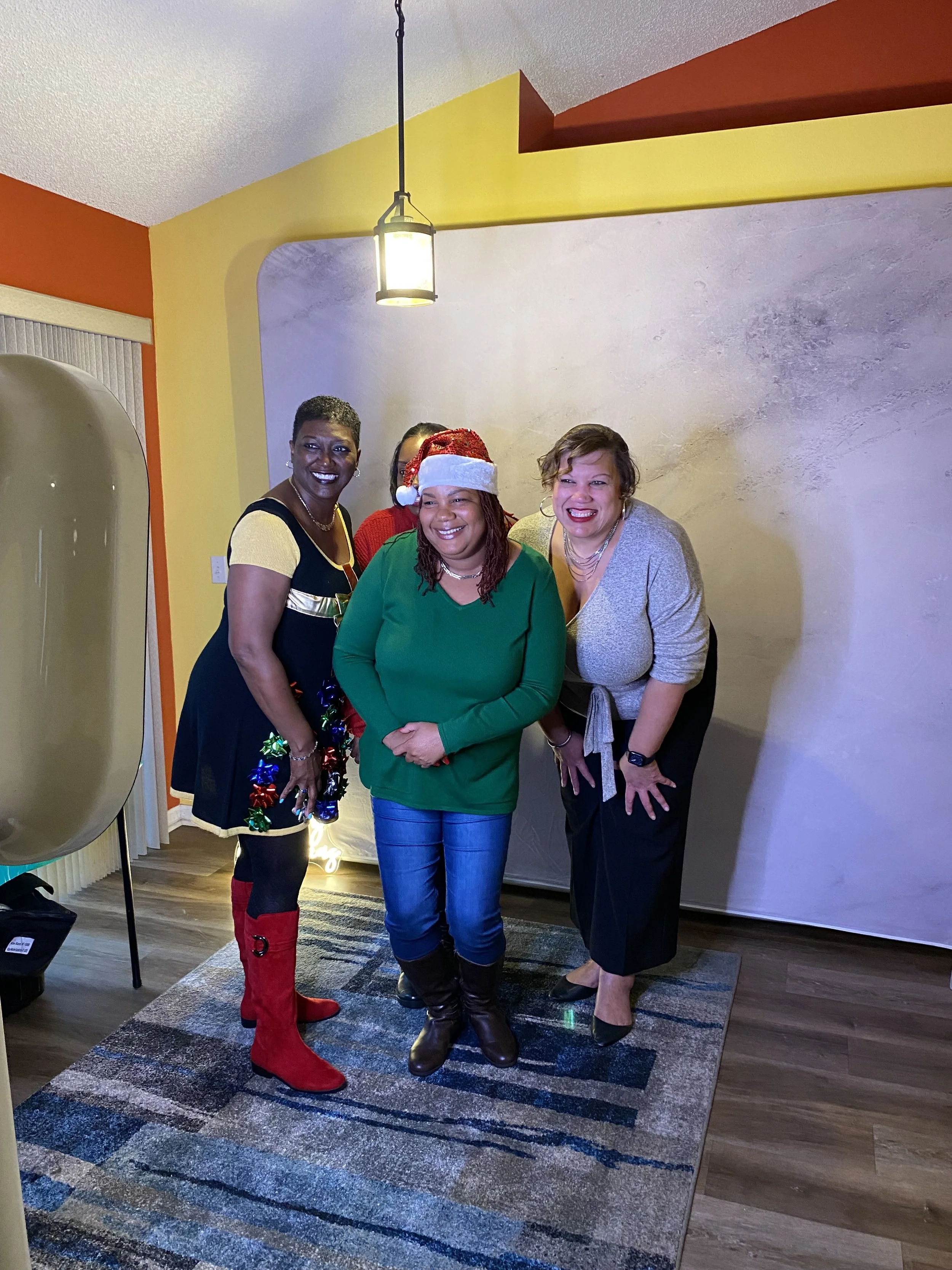 Four women smiling for a photo indoors during a holiday celebration, with one woman wearing a Santa hat and festive accessories.