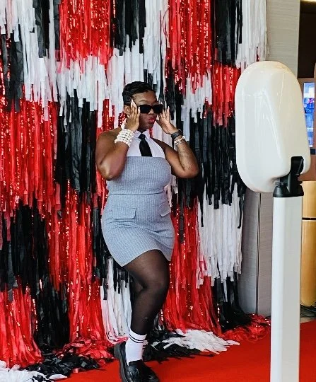 A woman posing in front of a red, black, and white decorative backdrop, wearing sunglasses, a gray sleeveless dress, black tights, and black shoes, with hair styled in an updo.