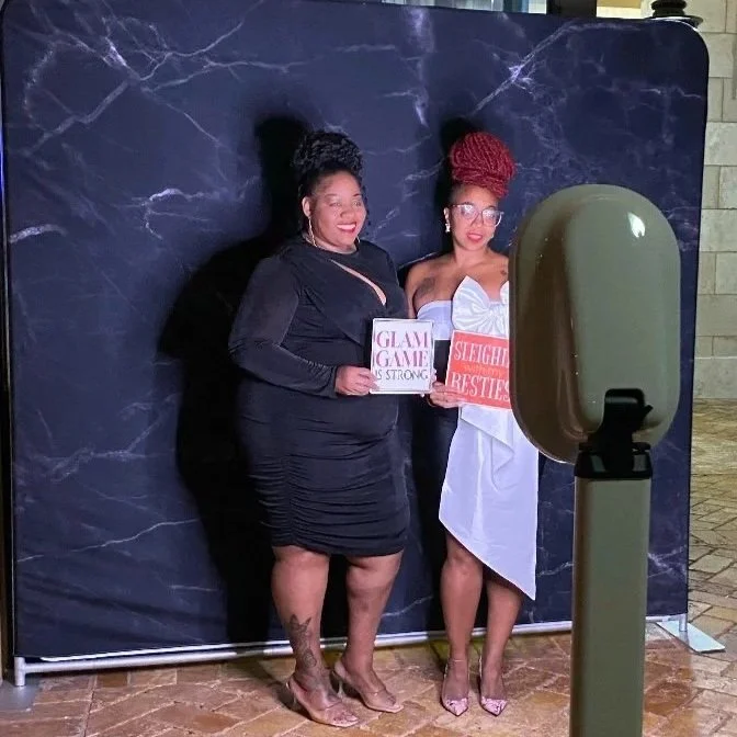 Two women standing together in front of a dark backdrop. One woman is wearing a black dress and holding a sign that says "Glam Game is Strong." The other woman is wearing a strapless white dress with a red sash that says "Sleighing the Besties" and i