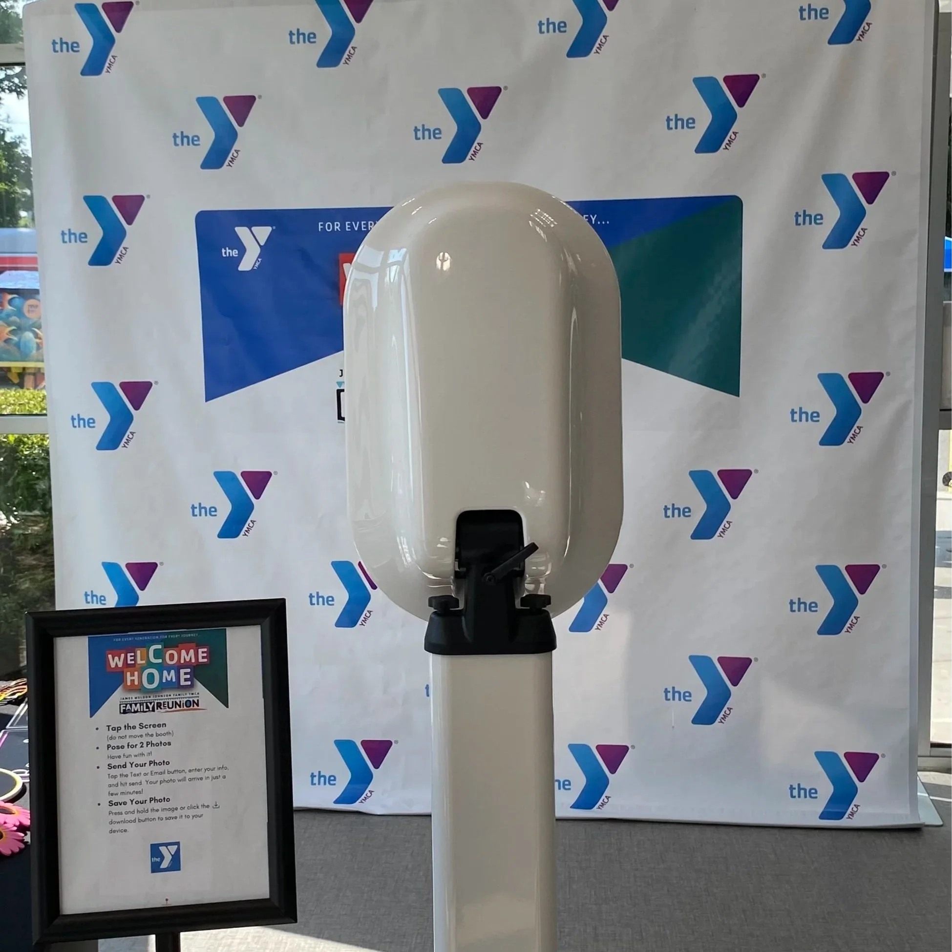 Photo of a photo booth with a YMCA background. The booth has instructions on how to take photos and a camera mounted on a stand for a family reunion event.