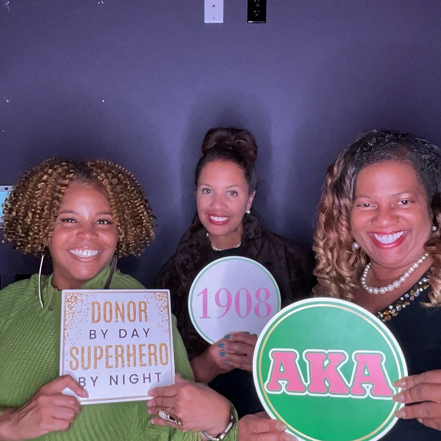 Three women smiling, holding signs and celebrating. The woman on the left is holding a sign that reads 'DONOR BY DAY SUPERHERO BY NIGHT'. The woman in the middle is holding a circular sign with the number '1908'. The woman on the right is holding a r