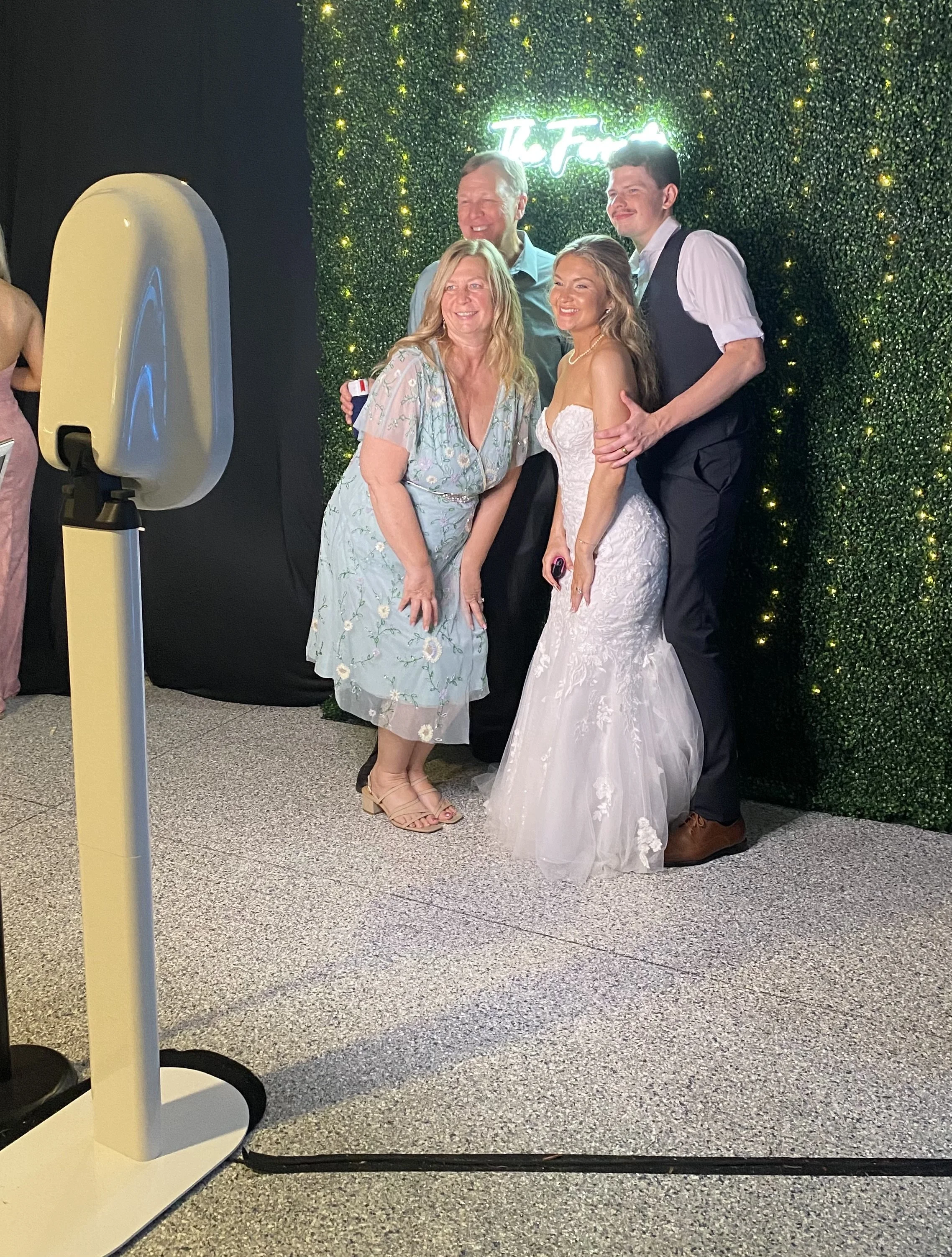 Four people are posing for a photo in front of a green garden wall with a neon sign. The group includes a woman in a floral dress, a woman in a white wedding dress, and two men dressed semi-formally. They are smiling and standing close together.