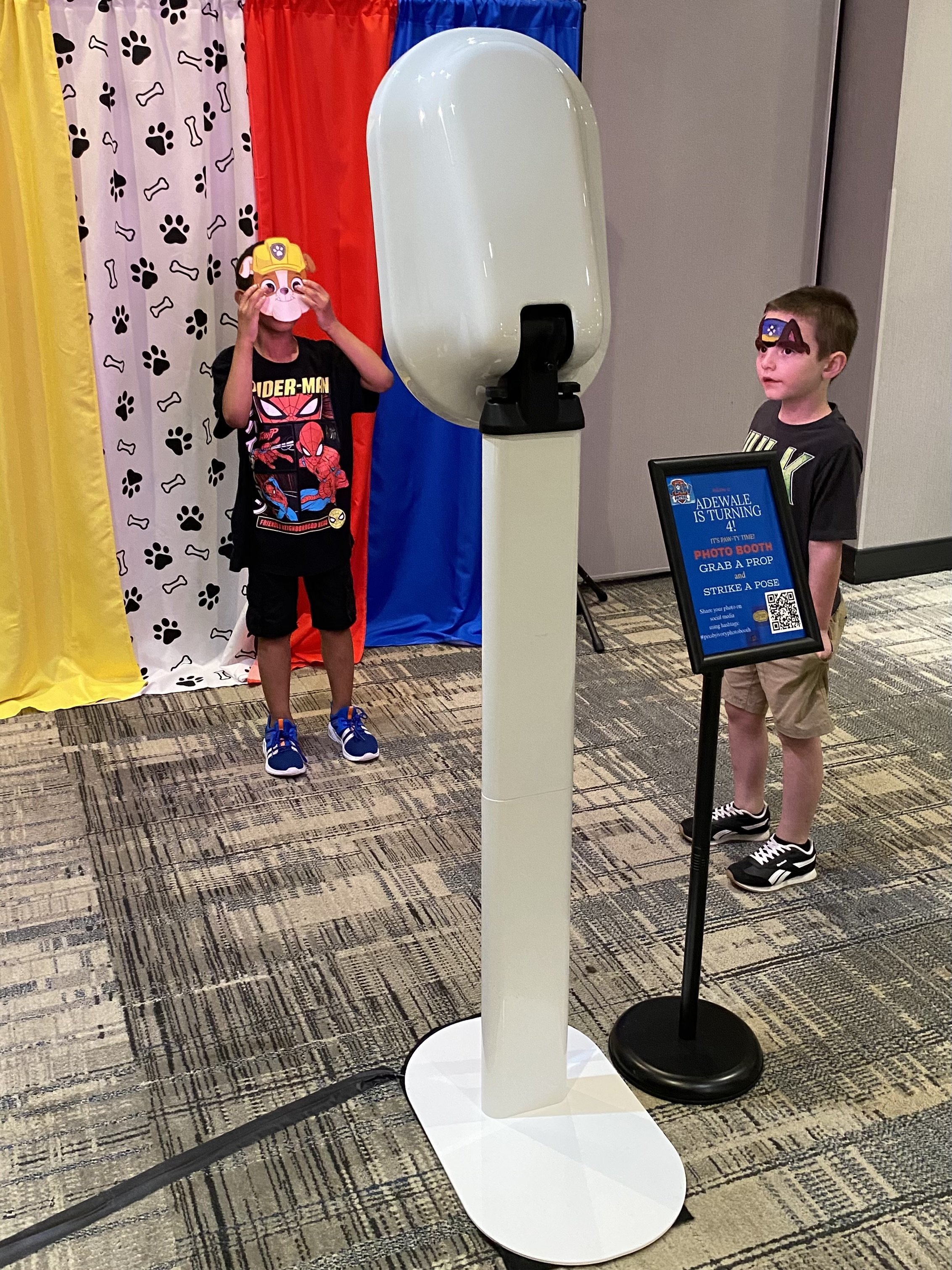 Two children at a photo booth with face masks, one wearing a Paw Patrol mask and the other a police dog mask, standing near a digital sign that says 'AdeWale is turning 4!', with a colorful curtain backdrop.